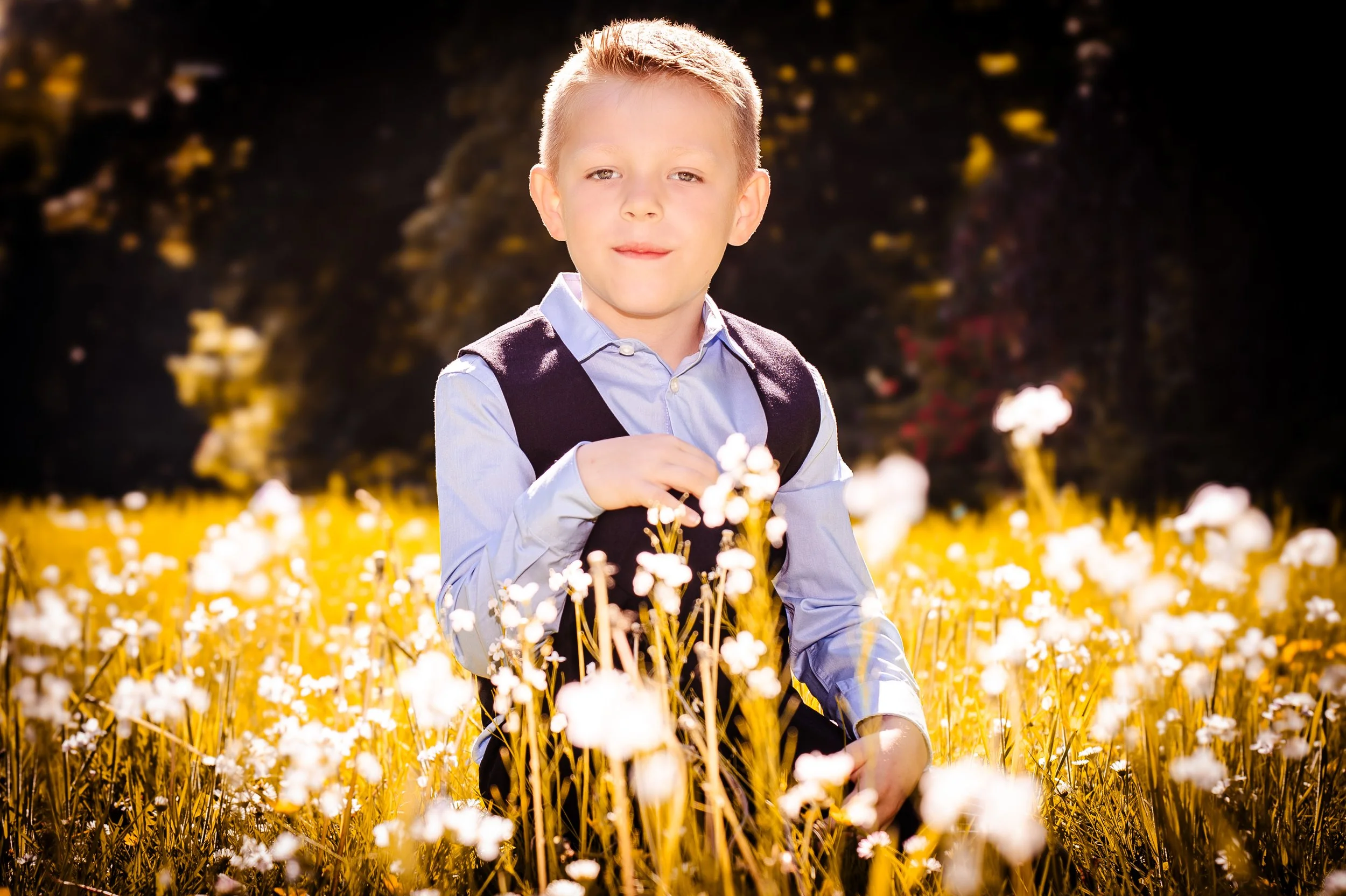 Jonge jongen in een veld vol witte bloemen, zittend en naar de camera kijkend, met een bosje bloemen in de hand op een zonnige dag.