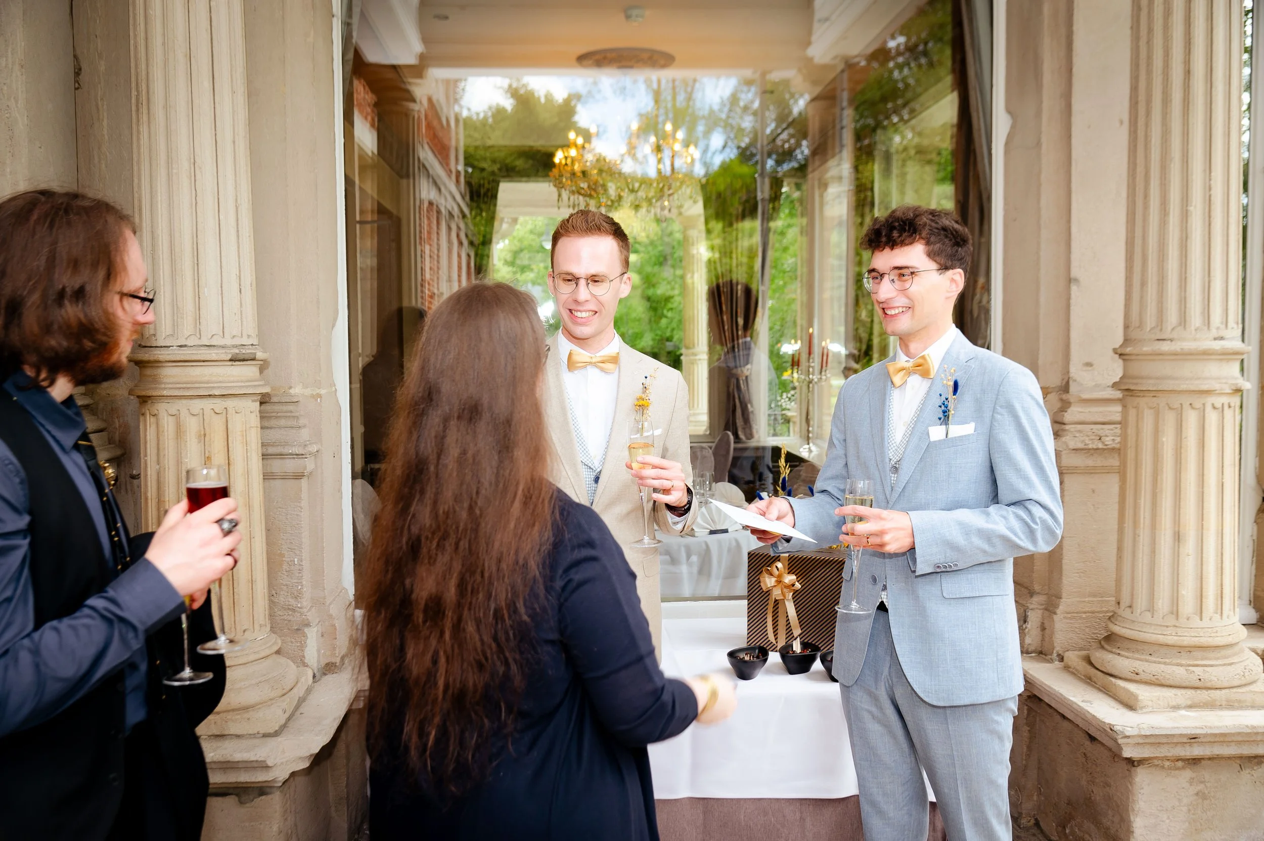 Jonge mannen en vrouw in formele kleding die toasten en praten bij een feestje, binnen en buiten een historisch gebouw met glazen muren en klassieke zuilen.