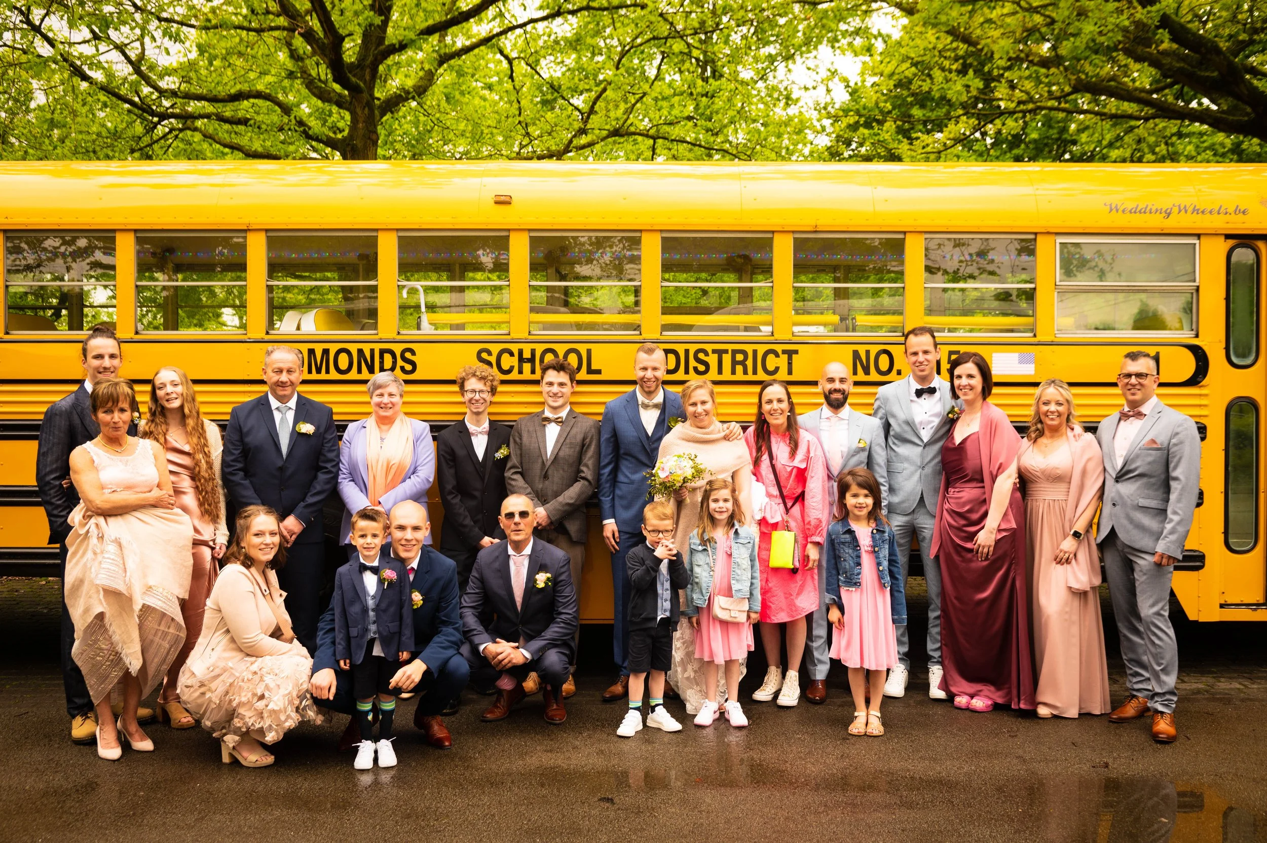 Groep mensen poseren voor een gele schoolbus met opschrift 'MONDS SCHOOL DISTRICT' tijdens een bruiloftsfeest, omgeven door groene bomen.