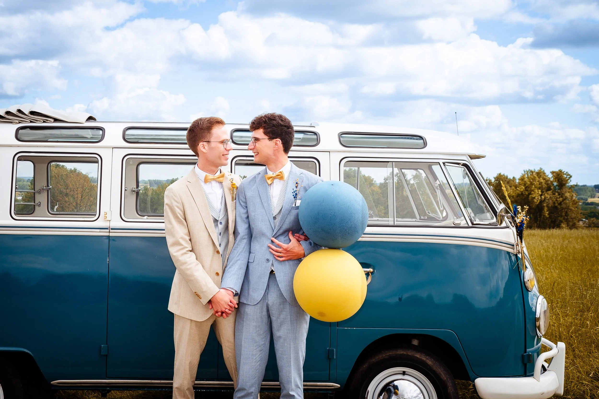 Twee mannen in gekleed in pastel kleurige pakken en gele boeketten op een veld, met een vintage blauwe bus op de achtergrond.