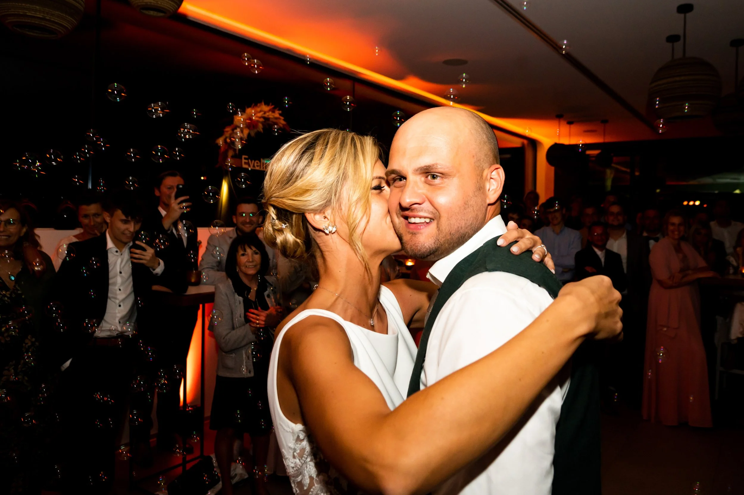Een bruid en bruidegom dansen samen op hun bruiloft met gasten op de achtergrond. De bruid kust de bruidegom op de wang.