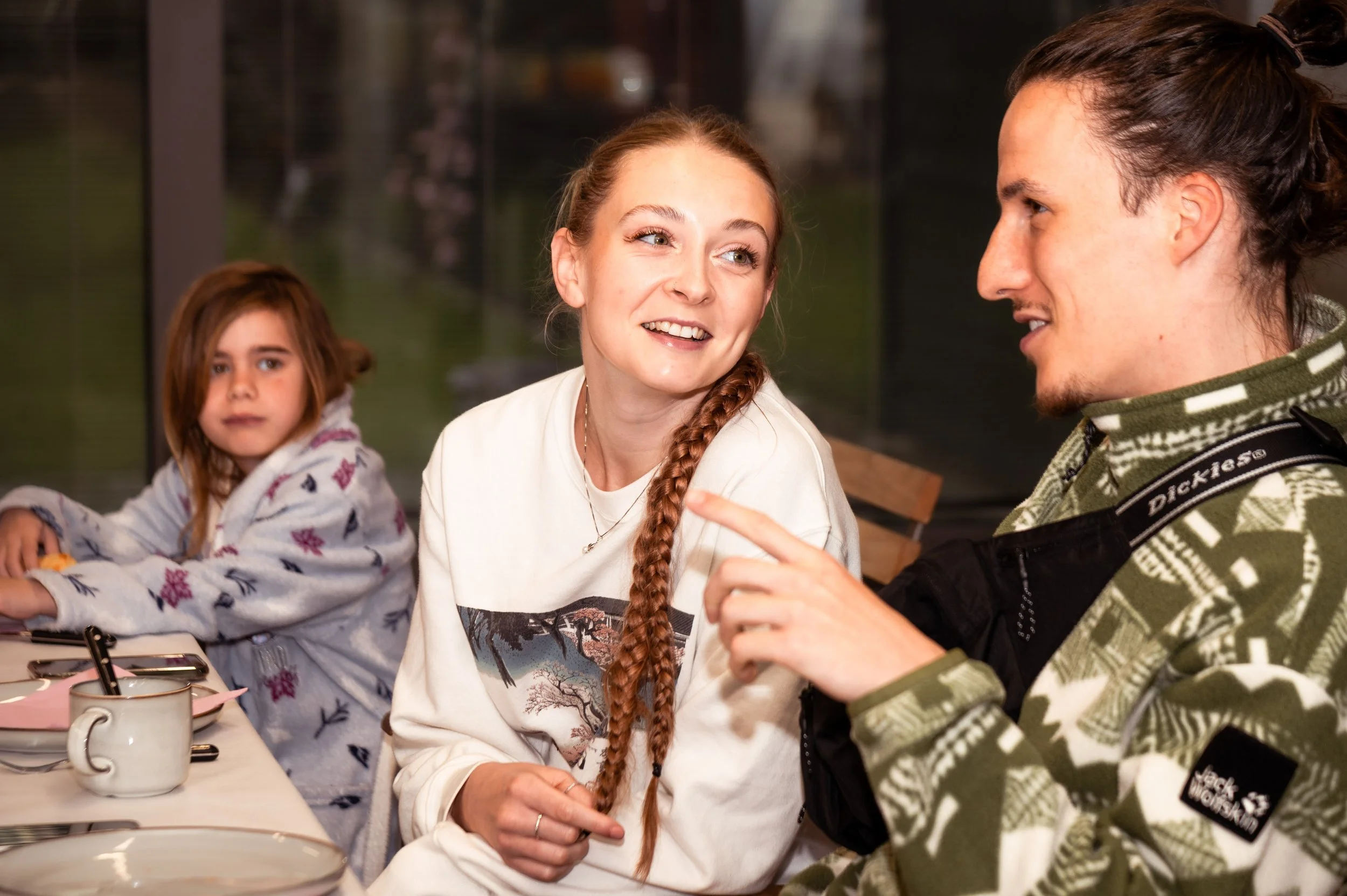 Drie mensen aan een tafel, een vrouw, een man en een jong meisje, die in gesprek zijn in een interieur, met glazen en bestek op tafel.