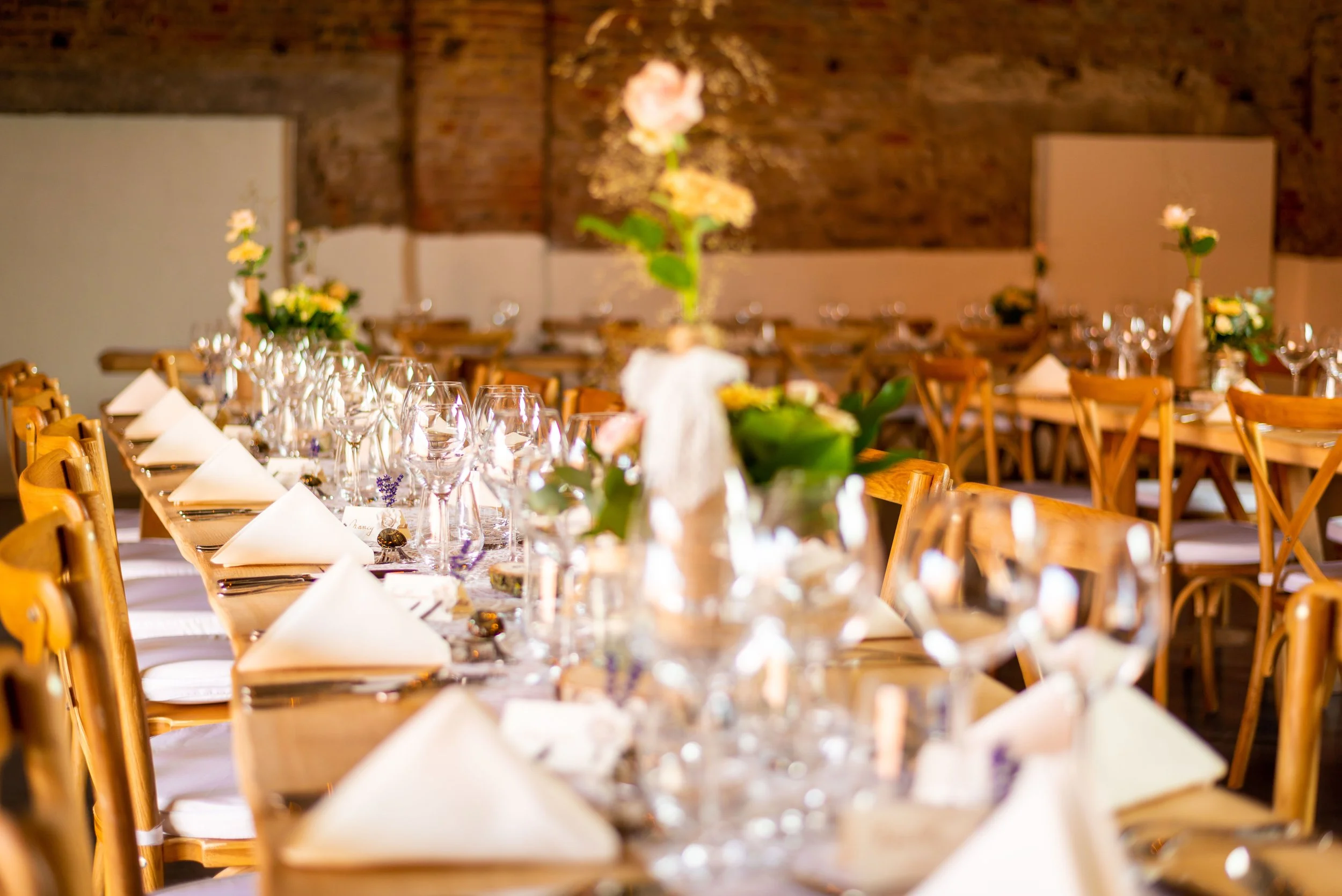 Feestzaal met lange gedekte tafels, witte servetten, wijn- en waterglazen, en bloemstukken, met een bakstenen muur op de achtergrond.