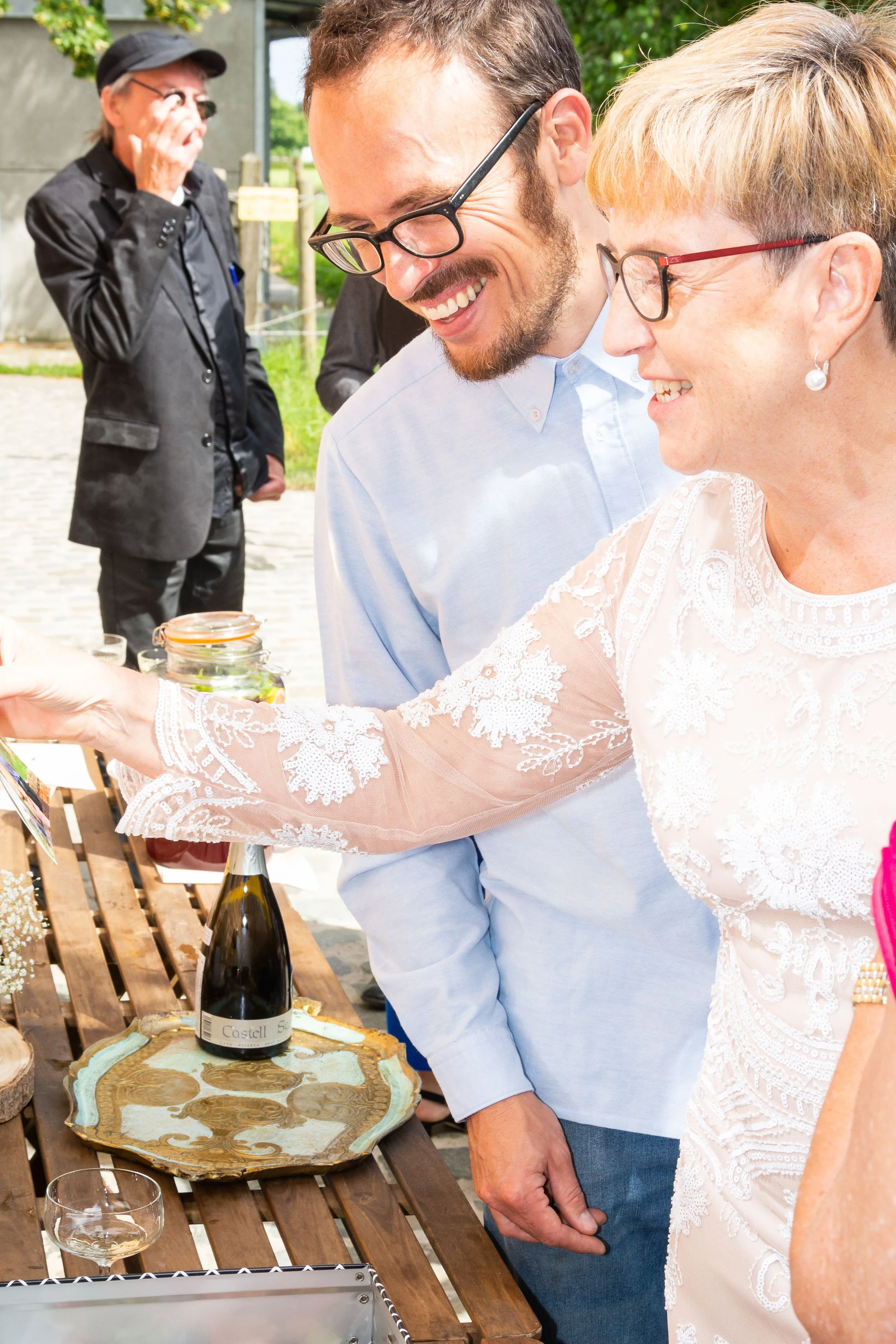Mensen die een fles champagne openen op een houten tafel tijdens een buitenfeest, met een man in een zwarte jas op de achtergrond.