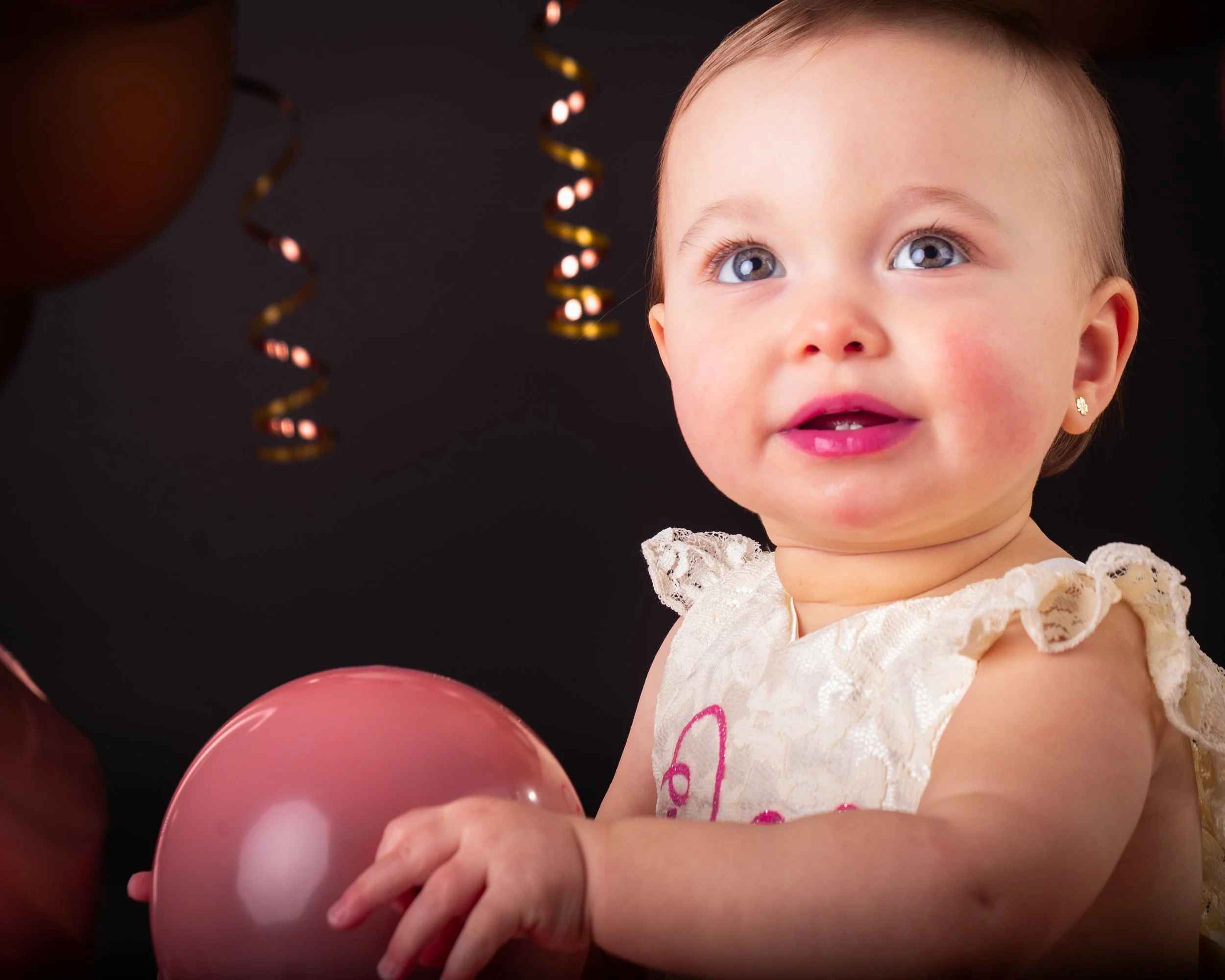 een fotos van een baby met blauwe ogen en een gebloemd jurkje die knuffels vasthoudt in een donkere kamer, met kleurrijke slingers op de achtergrond.