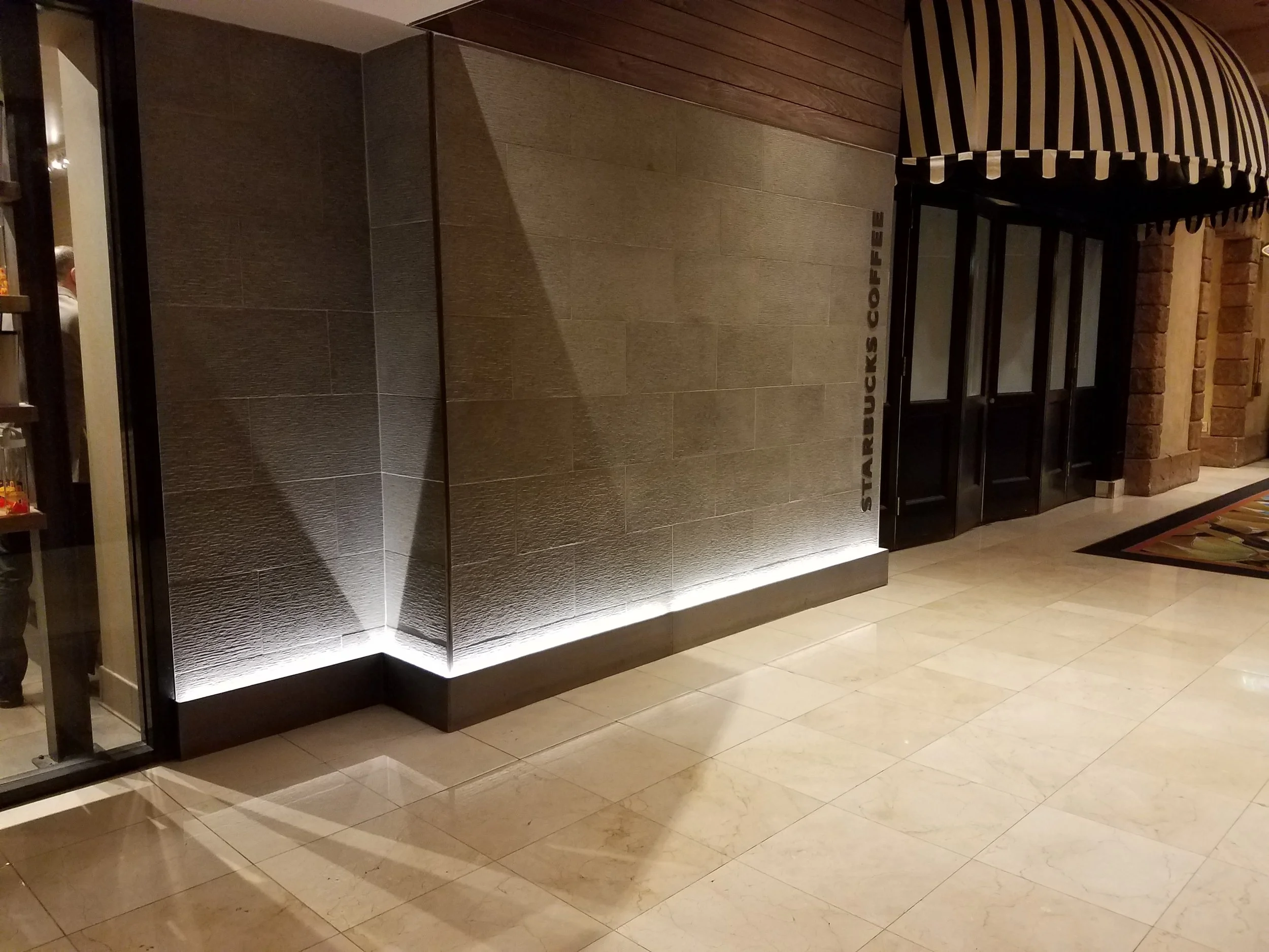 Entrance to a Starbucks Coffee store with a black awning featuring white light stripes and a sign that reads 'Starbucks Coffee' along the wall. The door is black and glass, with a marble floor and walls.