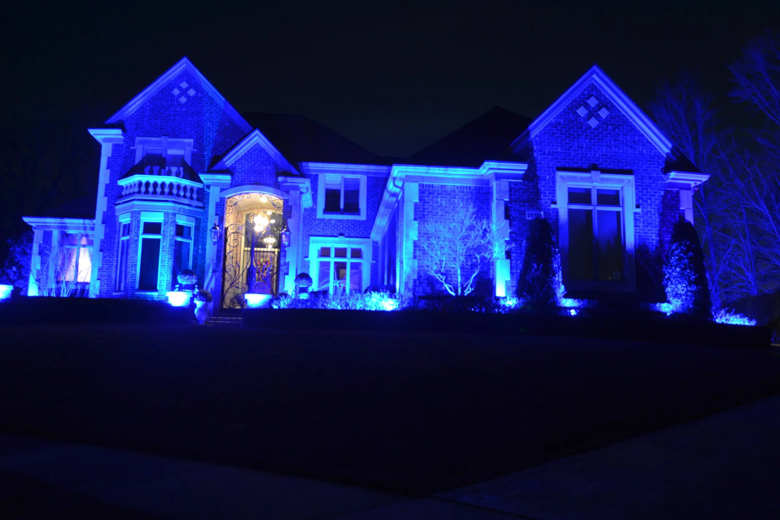 A large house illuminated with bright blue lights at night, showcasing its architectural features including multiple gables, bay windows, and a front entrance with decorative metalwork.