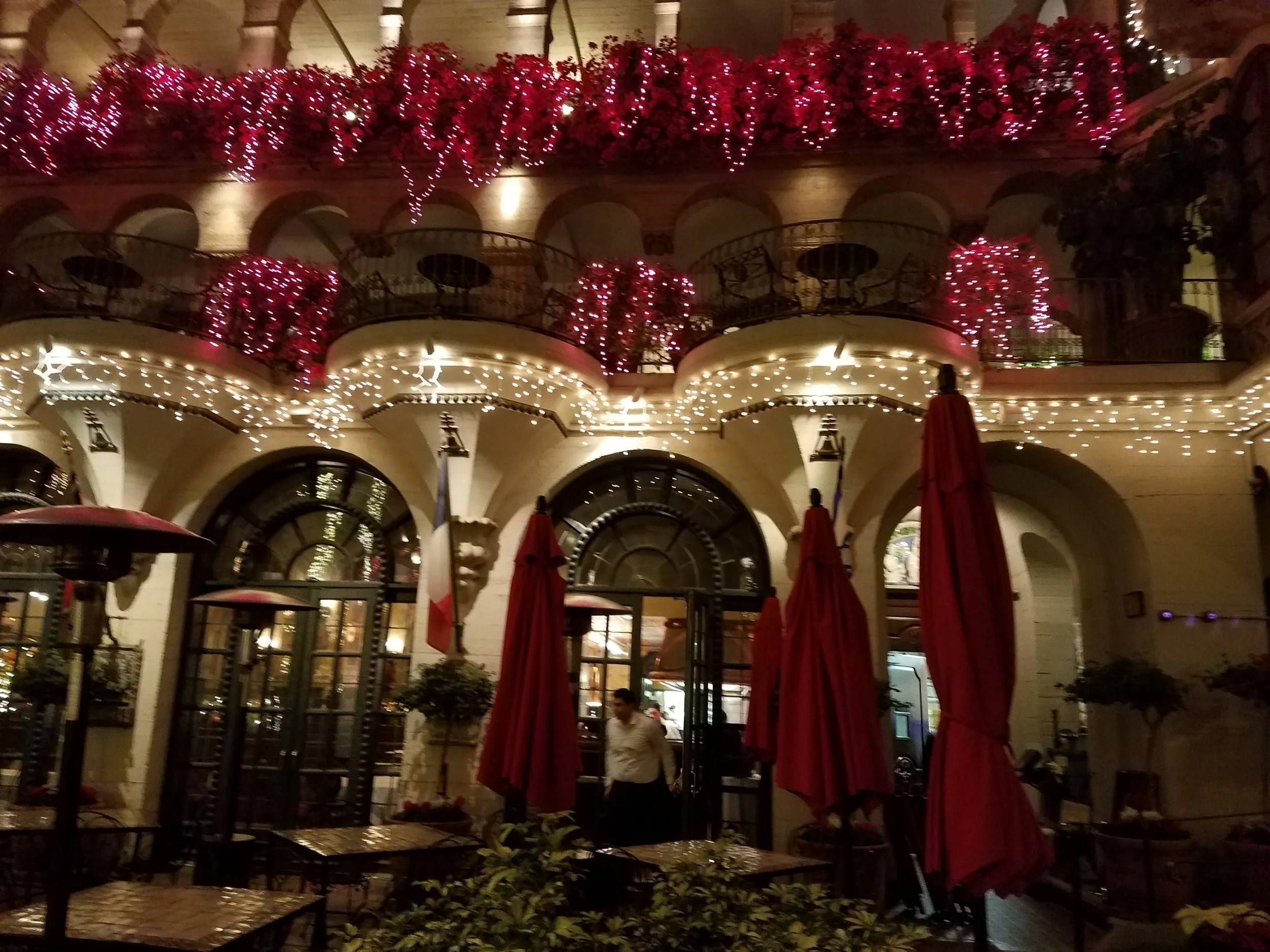 A multi-level building decorated with red flowers, string lights, and red umbrellas at night, with a restaurant entrance and a person walking inside.