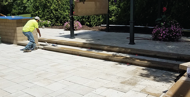 Construction worker installing paving stones on outdoor patio or walkway with flowers and greenery in the background.