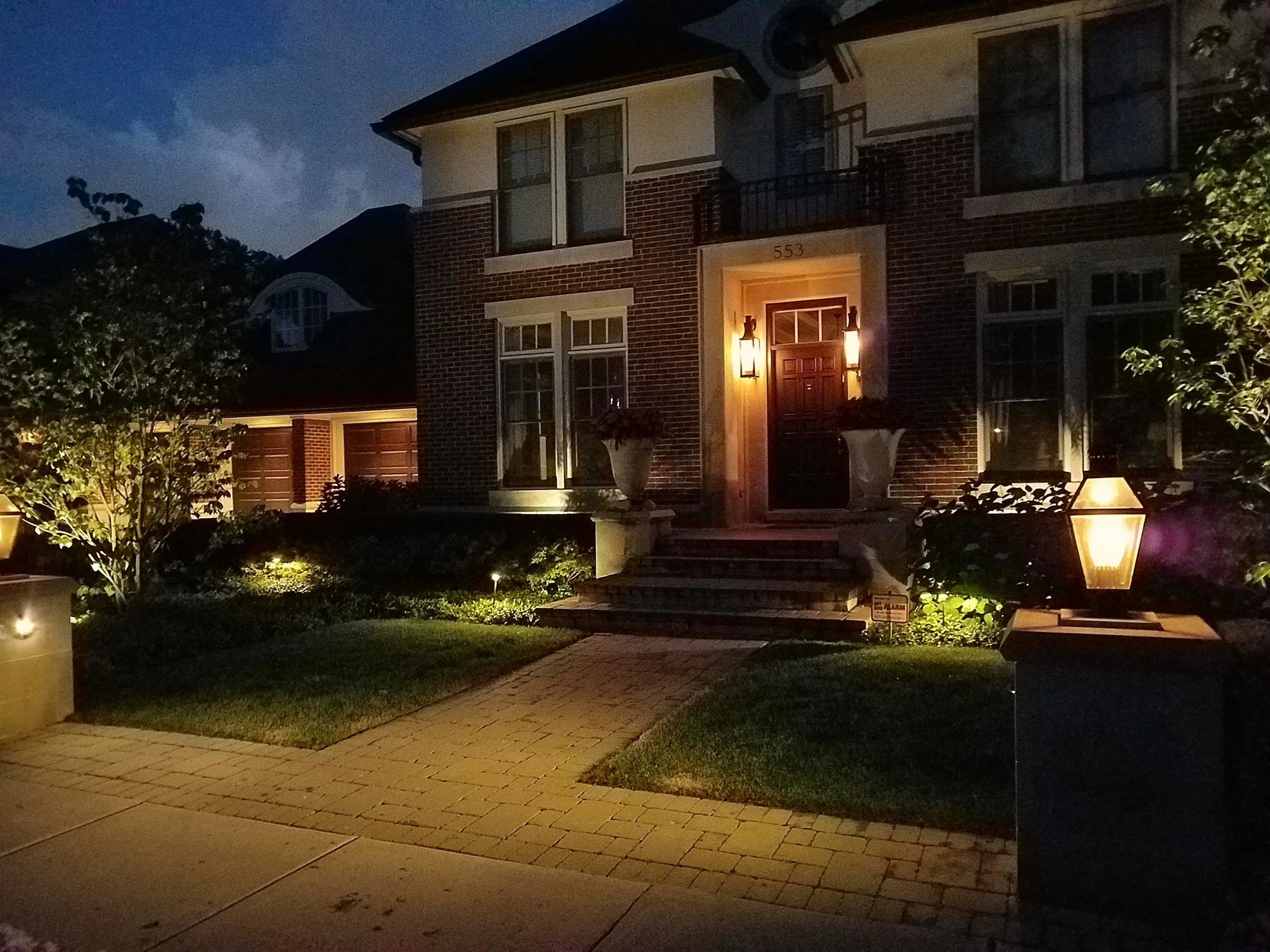 A large brick house at night with several porch lights illuminating the front yard and entryway. The entrance features steps, large potted plants, and a wooden door. The front yard has a well-maintained lawn, pathway, and garden lighting.