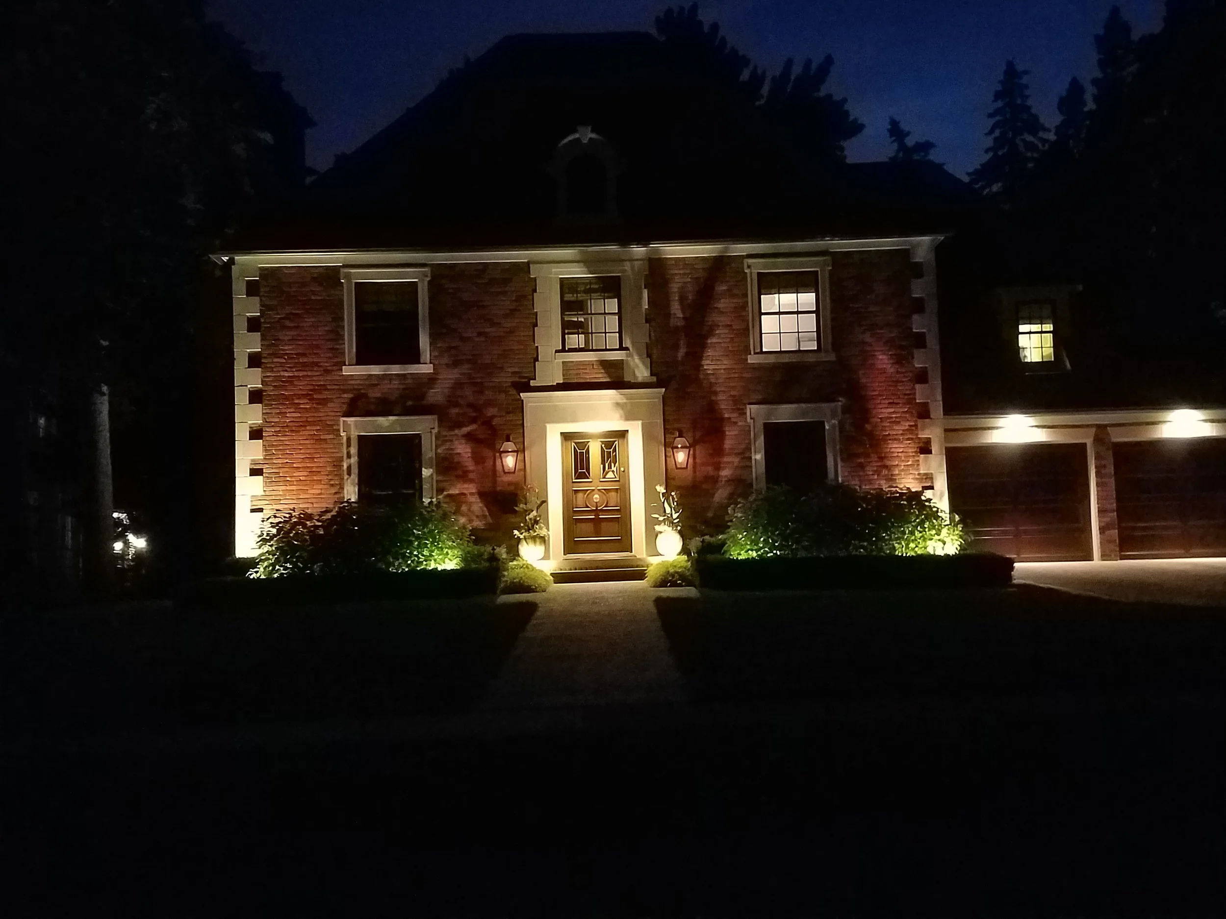 A two-story brick house illuminated by exterior lights at night, with a front door, windows, and surrounding landscaping.