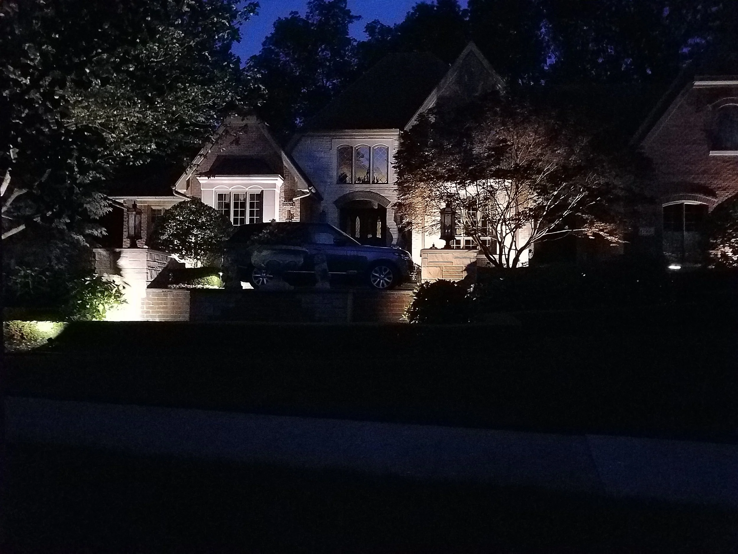 Nighttime image of a lit-up house with a driveway, parked car, trees, and shrubs.