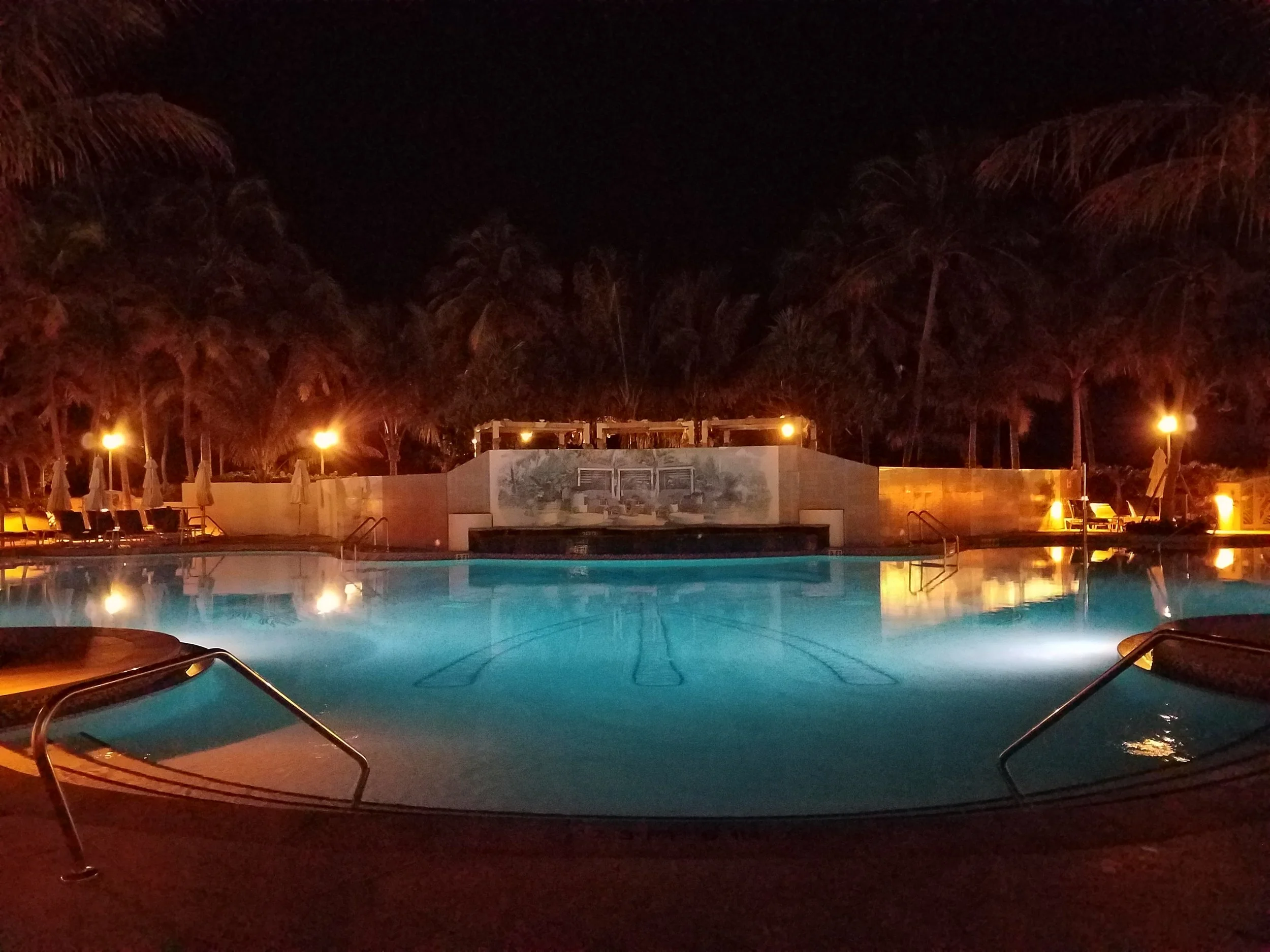 Empty illuminated swimming pool at night with palm trees and lounge chairs in the background.