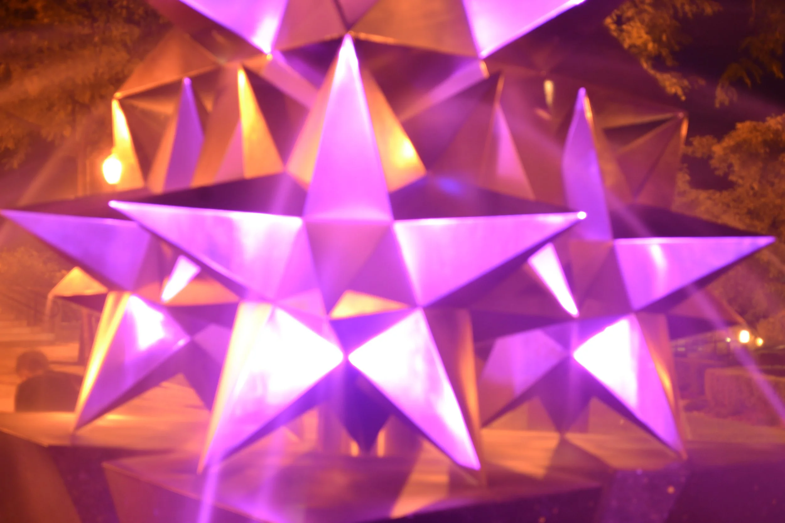 Large purple and pink illuminated star-shaped Christmas lantern at night with trees and a person in the background.