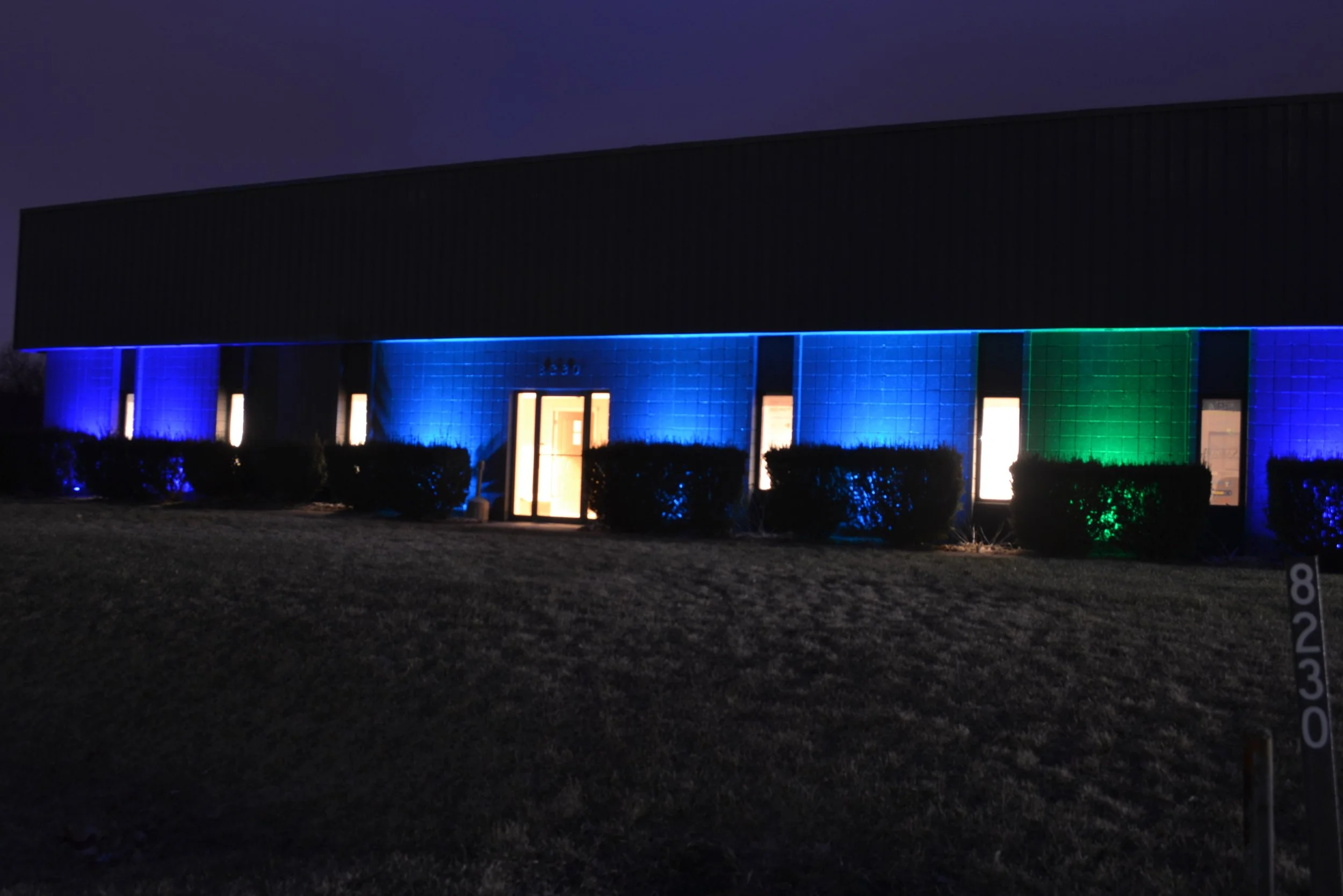 A modern building illuminated with blue and green lights at night, with bushes in front and a grassy area.