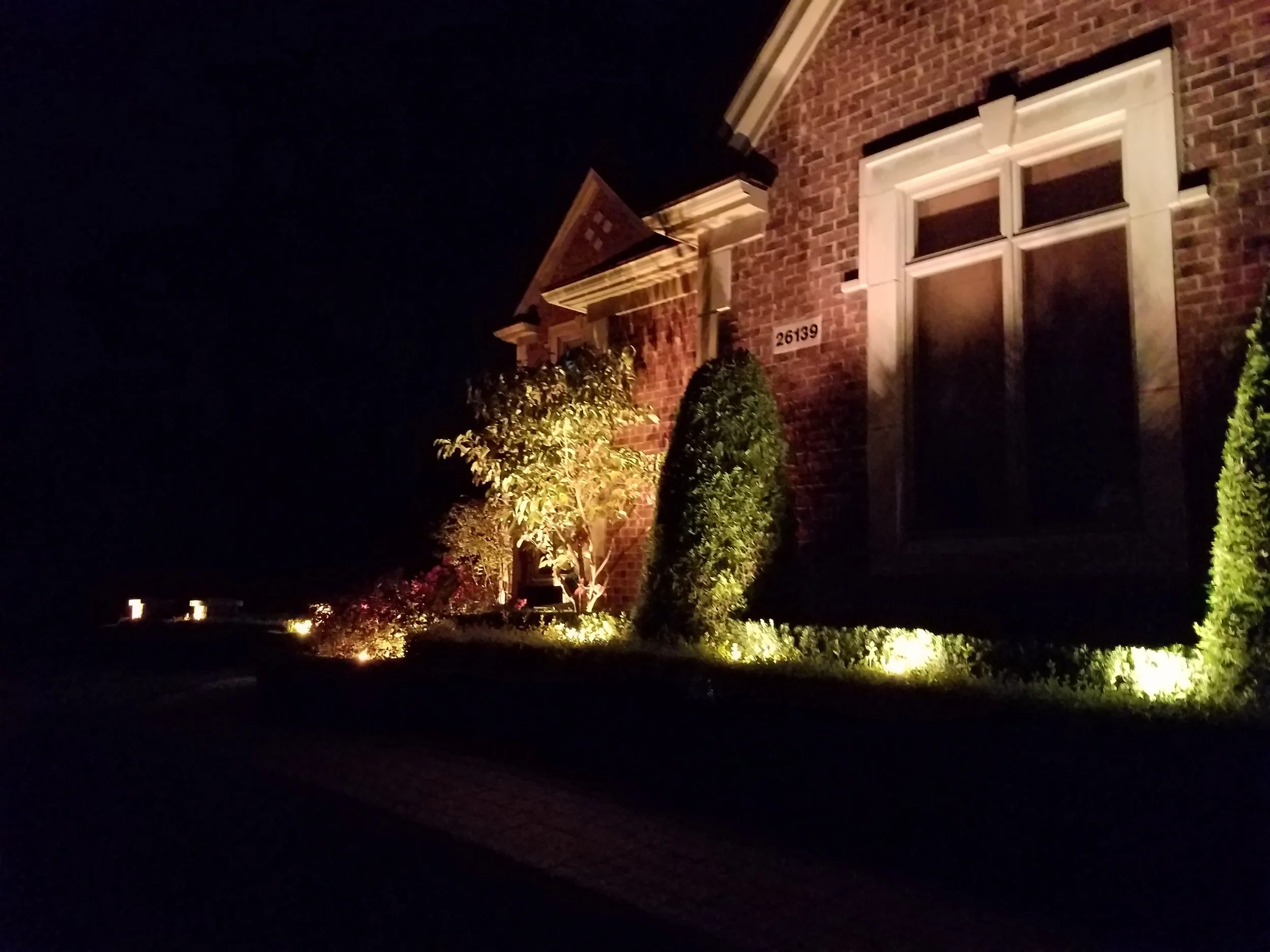 Night view of a brick house with illuminated landscaping including bushes and small trees.