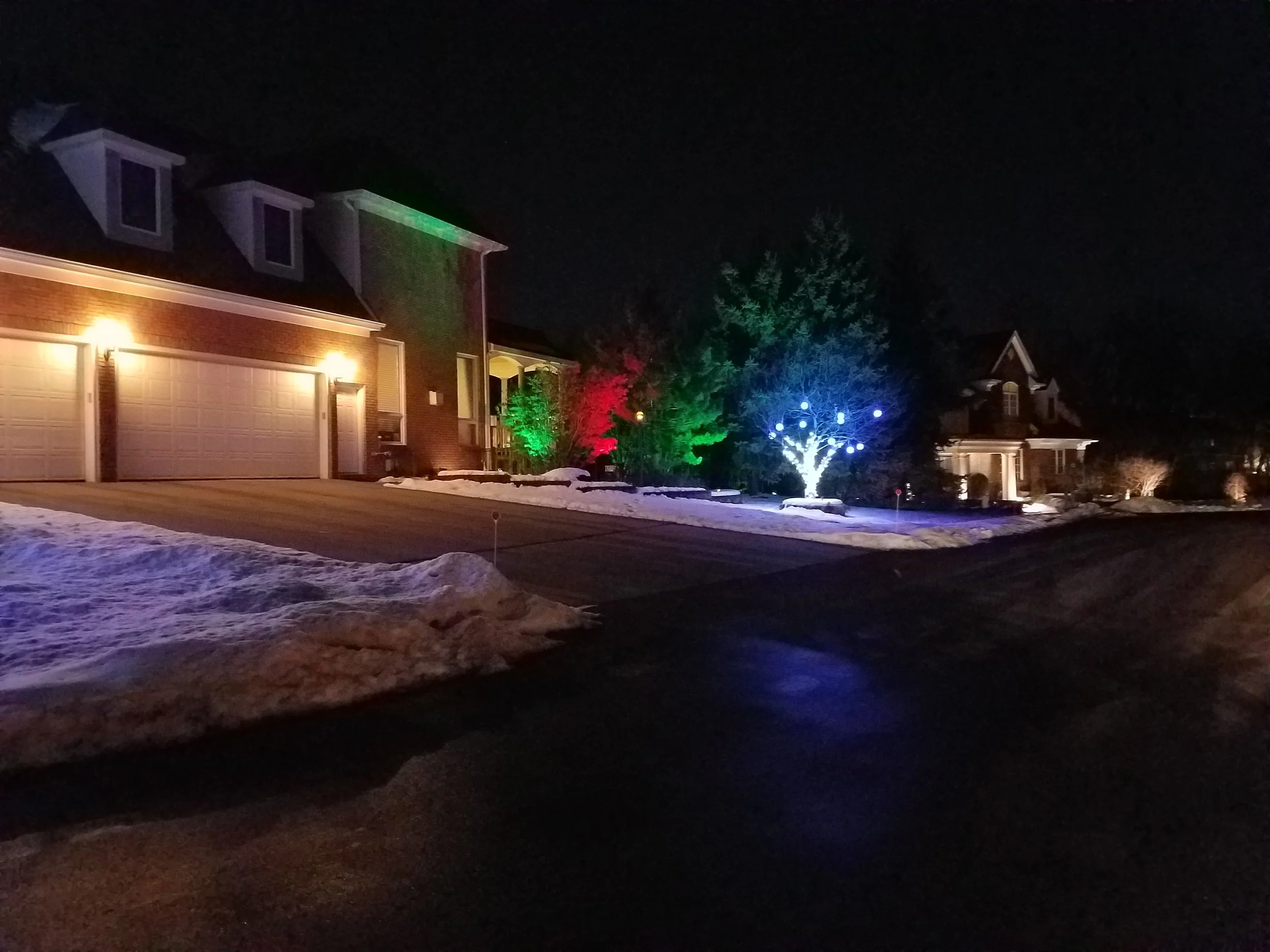Night view of a suburban neighborhood with houses and trees decorated with colorful Christmas lights, and snow on the ground.