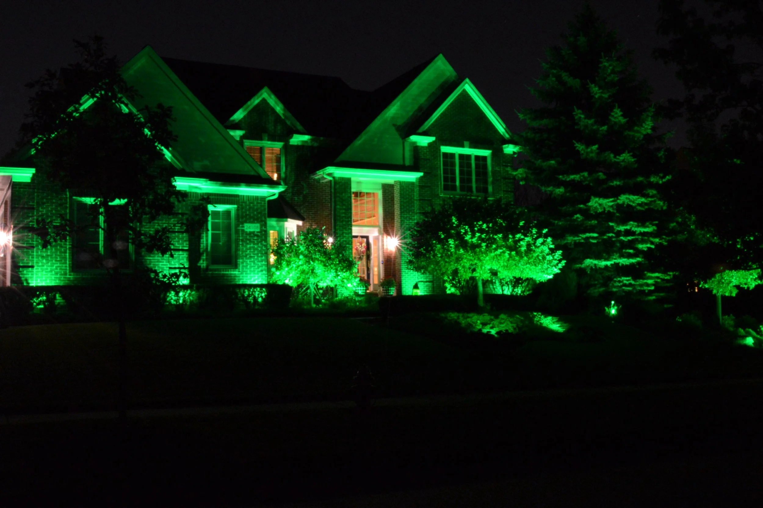 A house illuminated with green exterior lights at night, surrounded by trees and bushes.