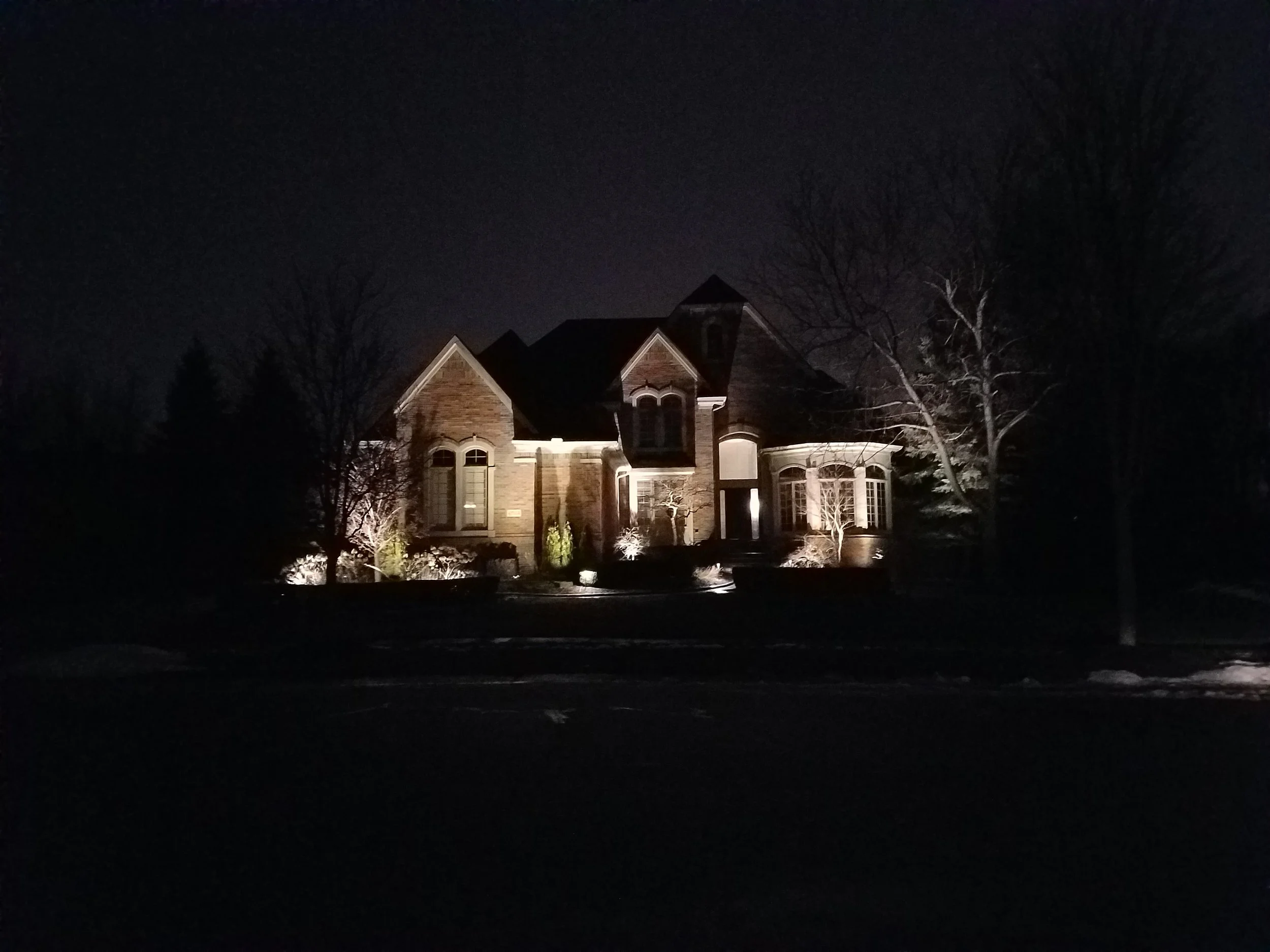 A large brick house with multiple gabled roofs, lit by exterior lights, at night. There are leafless trees surrounding the house and a dark street in the foreground.