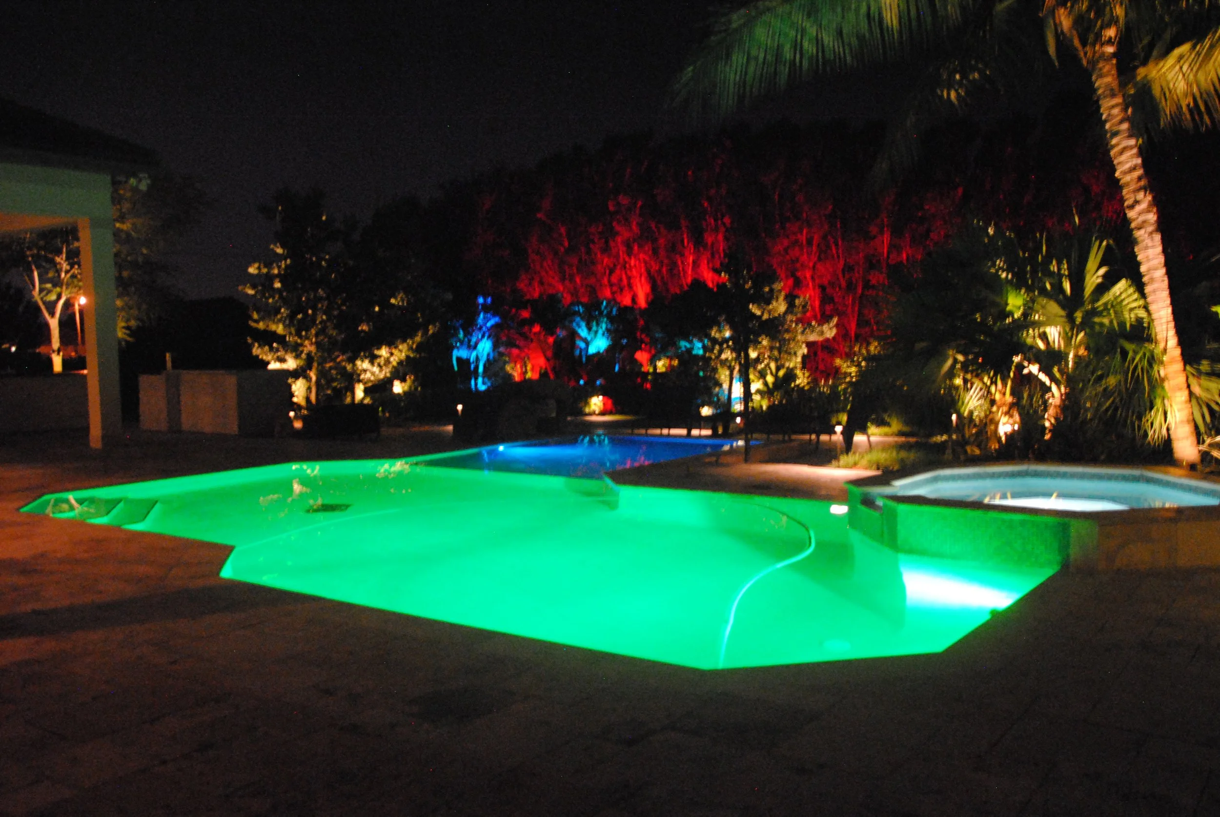 Night view of a swimming pool with colorful underwater lights, surrounded by trees and tropical plants, with a hot tub in the foreground.