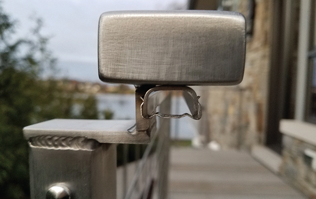 Close-up of a metallic balcony railing with a rectangular post and a glass panel holder, outdoors with a blurred house and trees in the background.