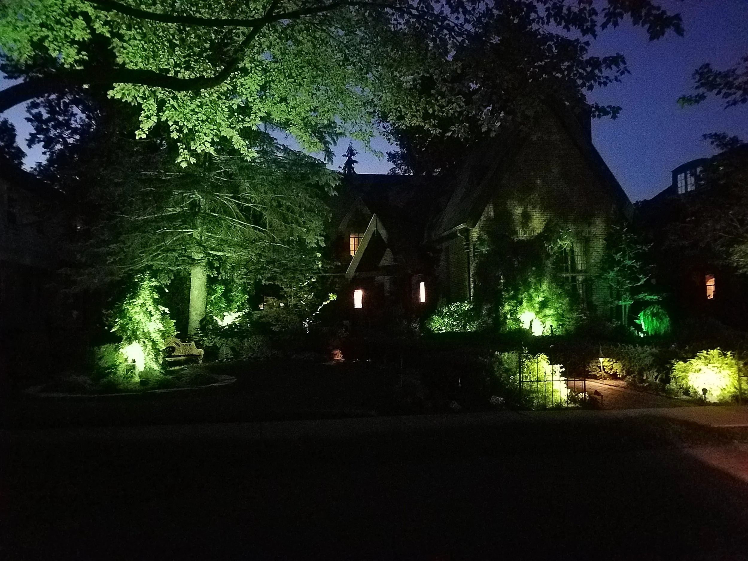 Nighttime scene of a house with trees illuminated by outdoor lighting, some windows glowing warmly.