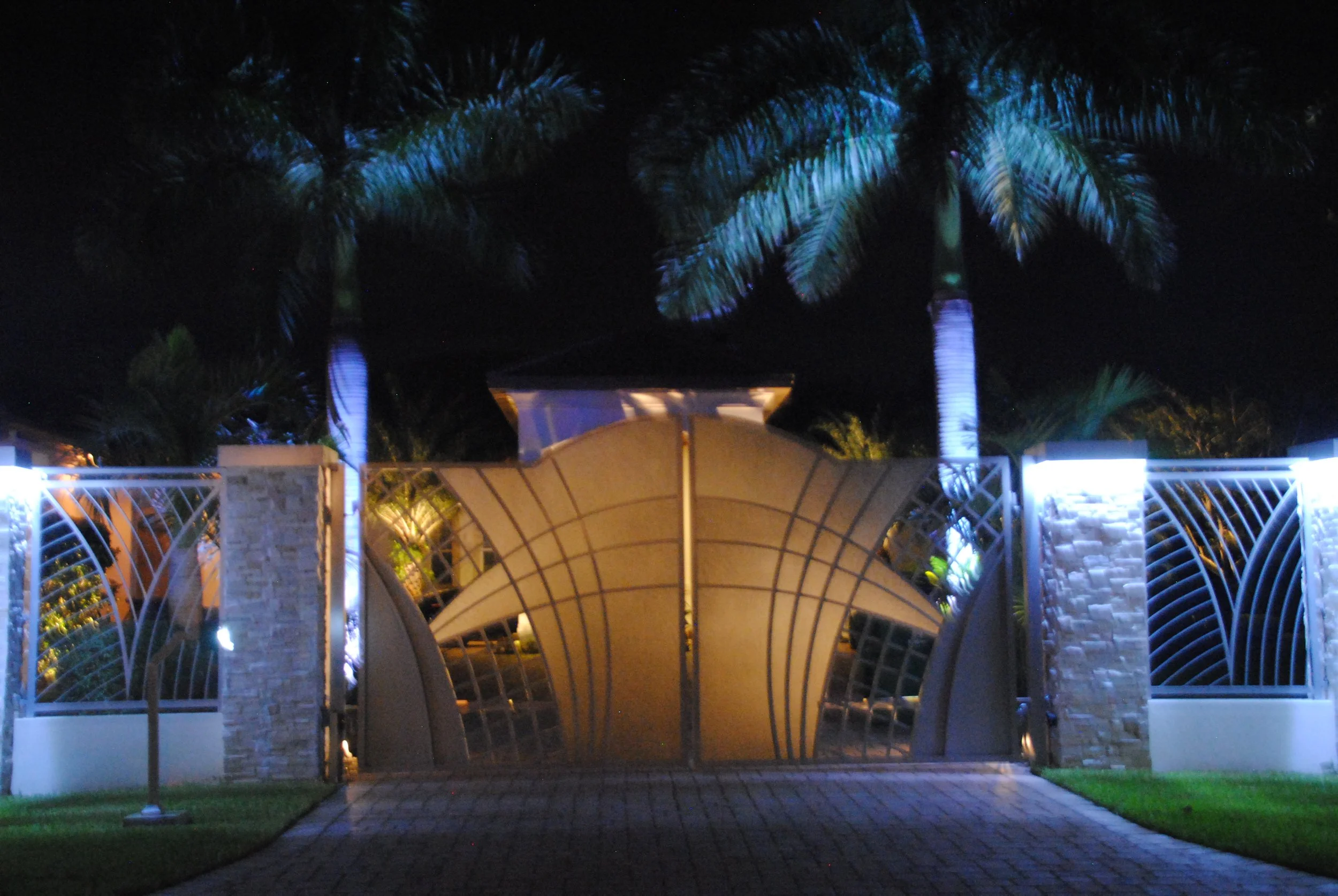 Night view of an ornate metal gate with two tall palm trees illuminated by blue and white lights, and a brick-paved driveway in the foreground.