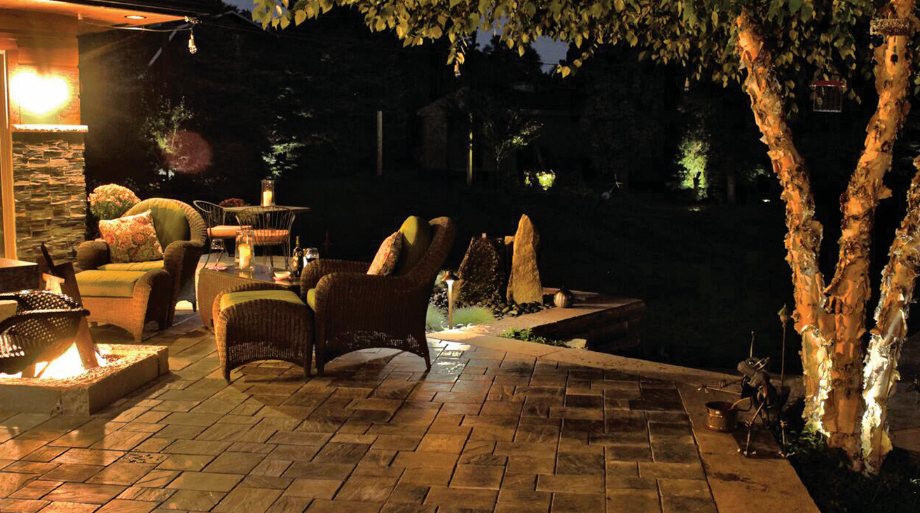 Nighttime outdoor patio with wicker and upholstered armchairs, small tables with candles, and a fire pit, surrounded by trees and illuminated landscaping.