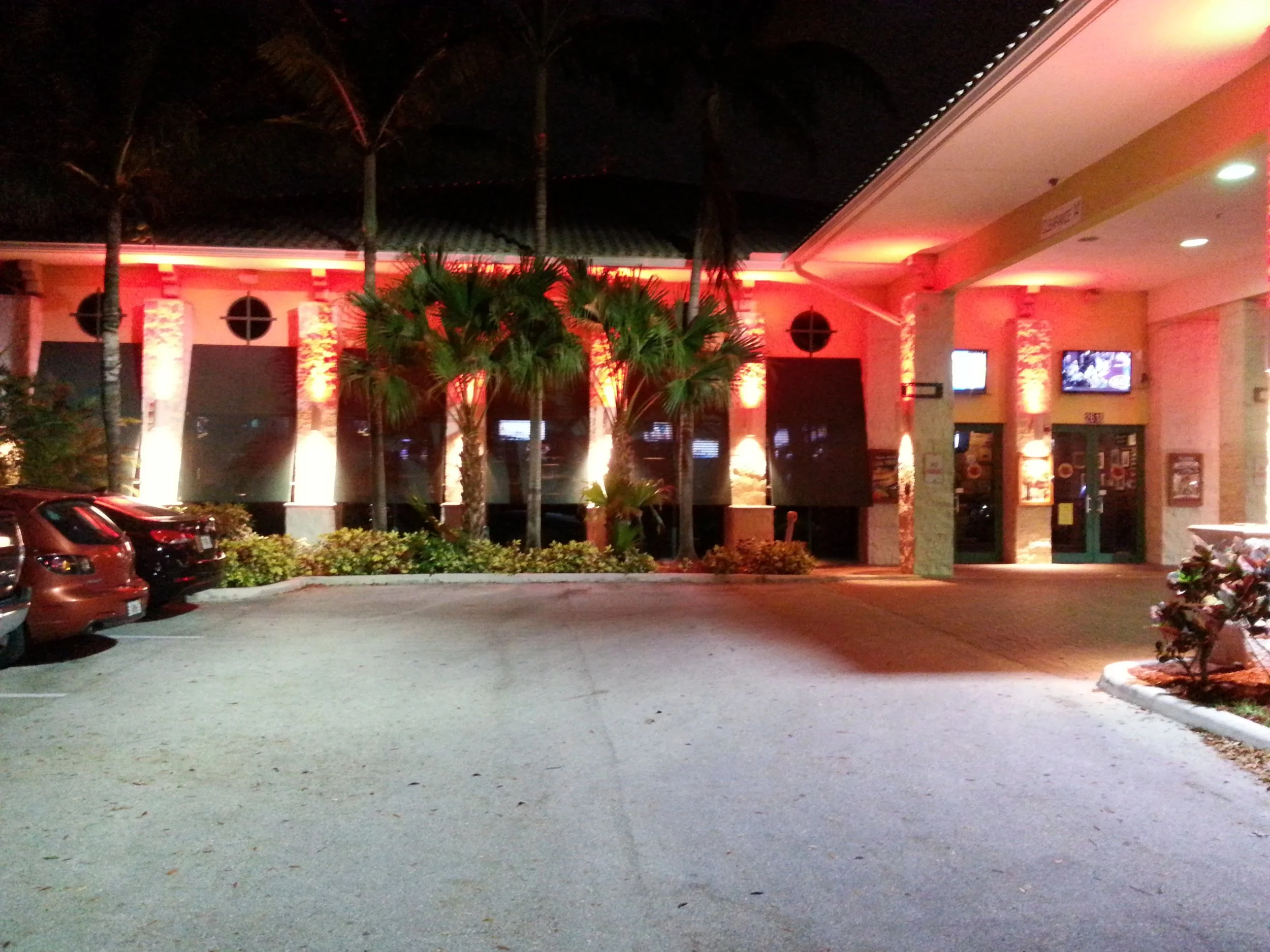 Night view of a well-lit restaurant or café with palm trees, cars parked in front, and a patio, illuminated with red and yellow lights.