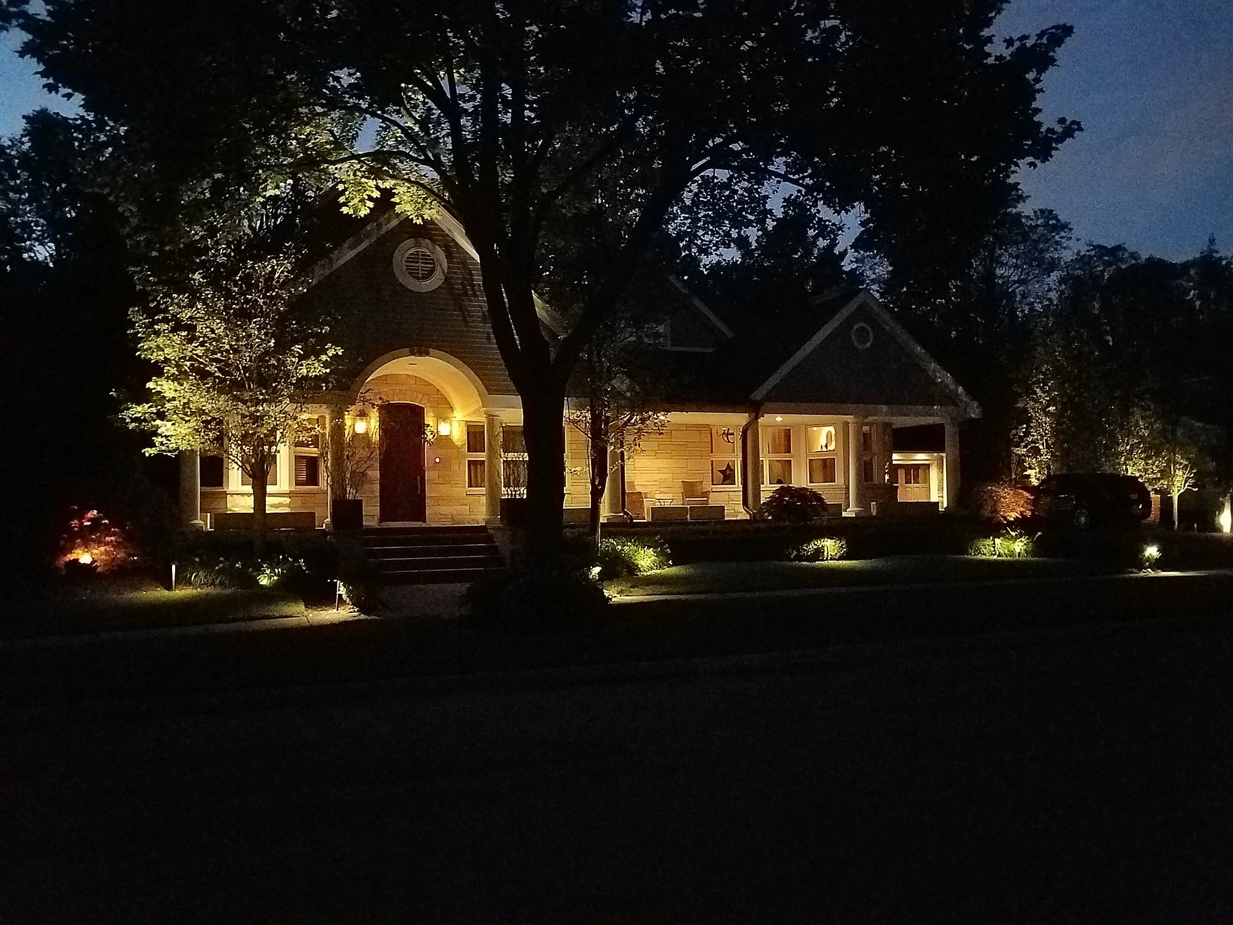 A house illuminated with exterior lights at night, surrounded by trees and landscaping.