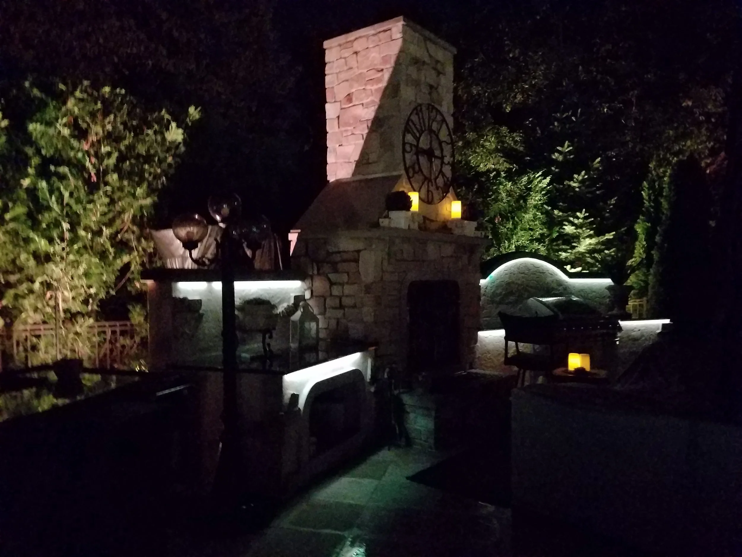Nighttime outdoor scene with a stone fireplace, a clock on the chimney, candles, and outdoor furniture illuminated by subtle lighting, surrounded by trees.