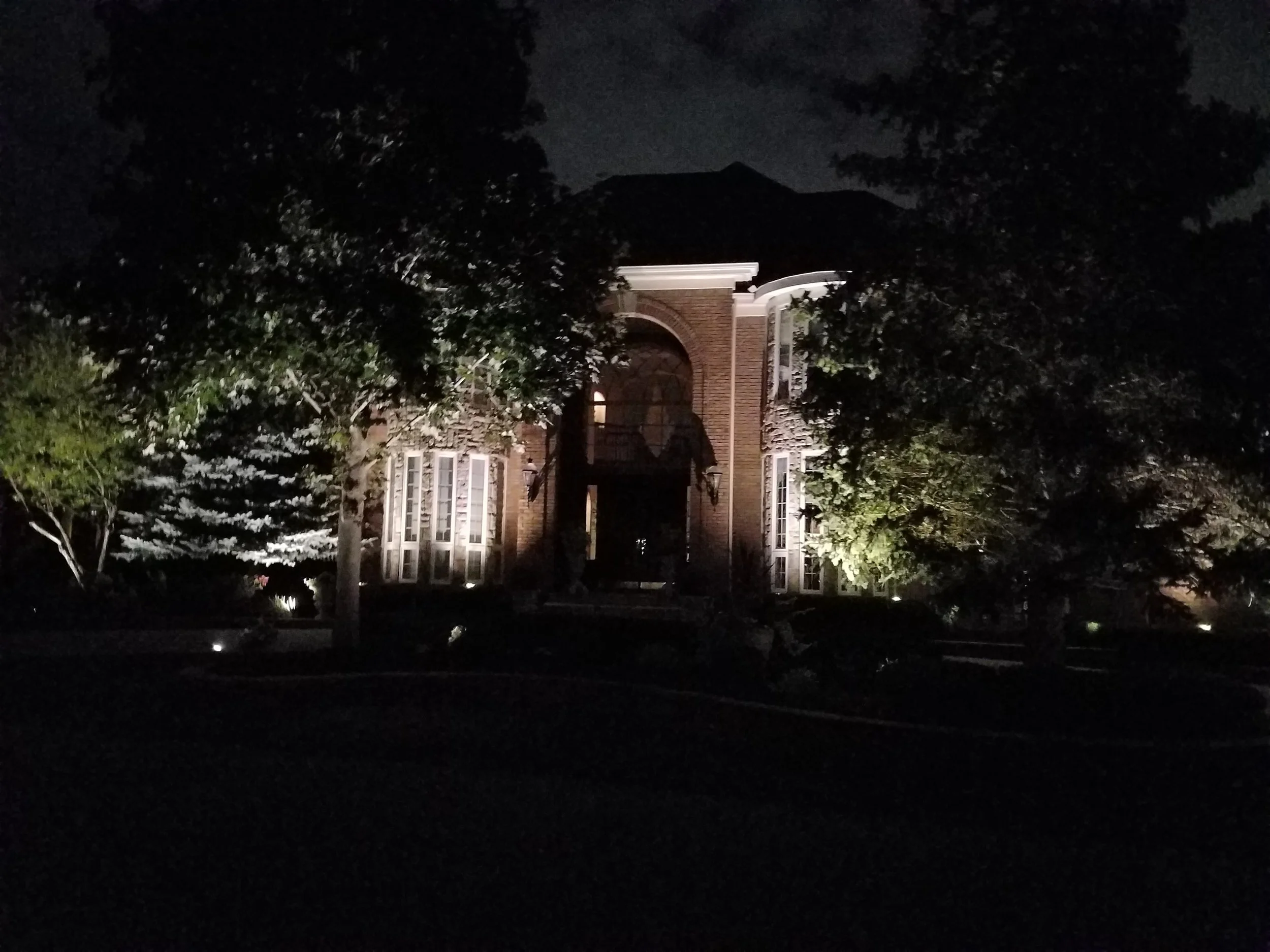 Nighttime view of a house with brick exterior, large windows, and trees in front, illuminated by exterior lights.