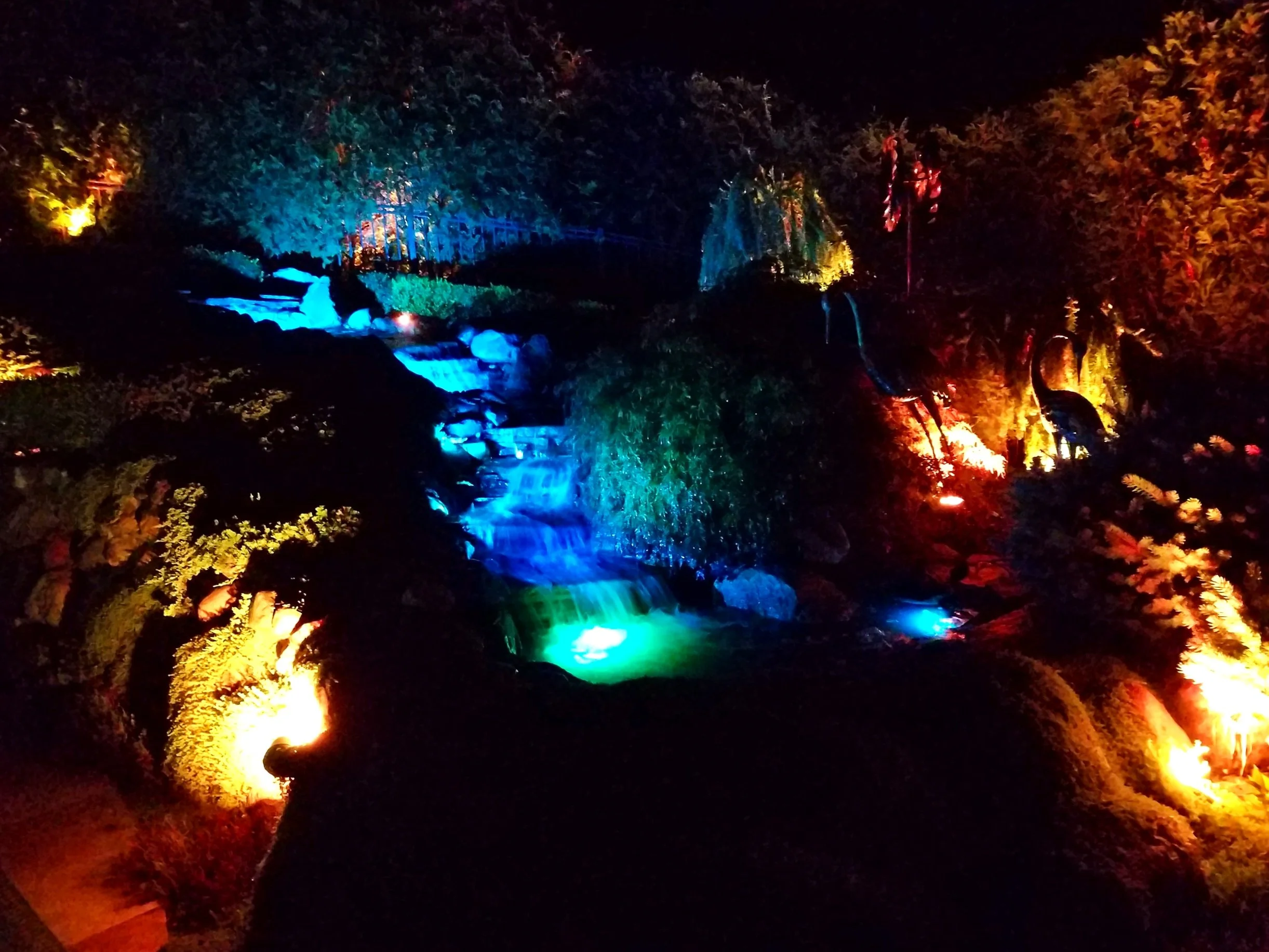 Nighttime colorful illuminated waterfall and crane statues in a garden.