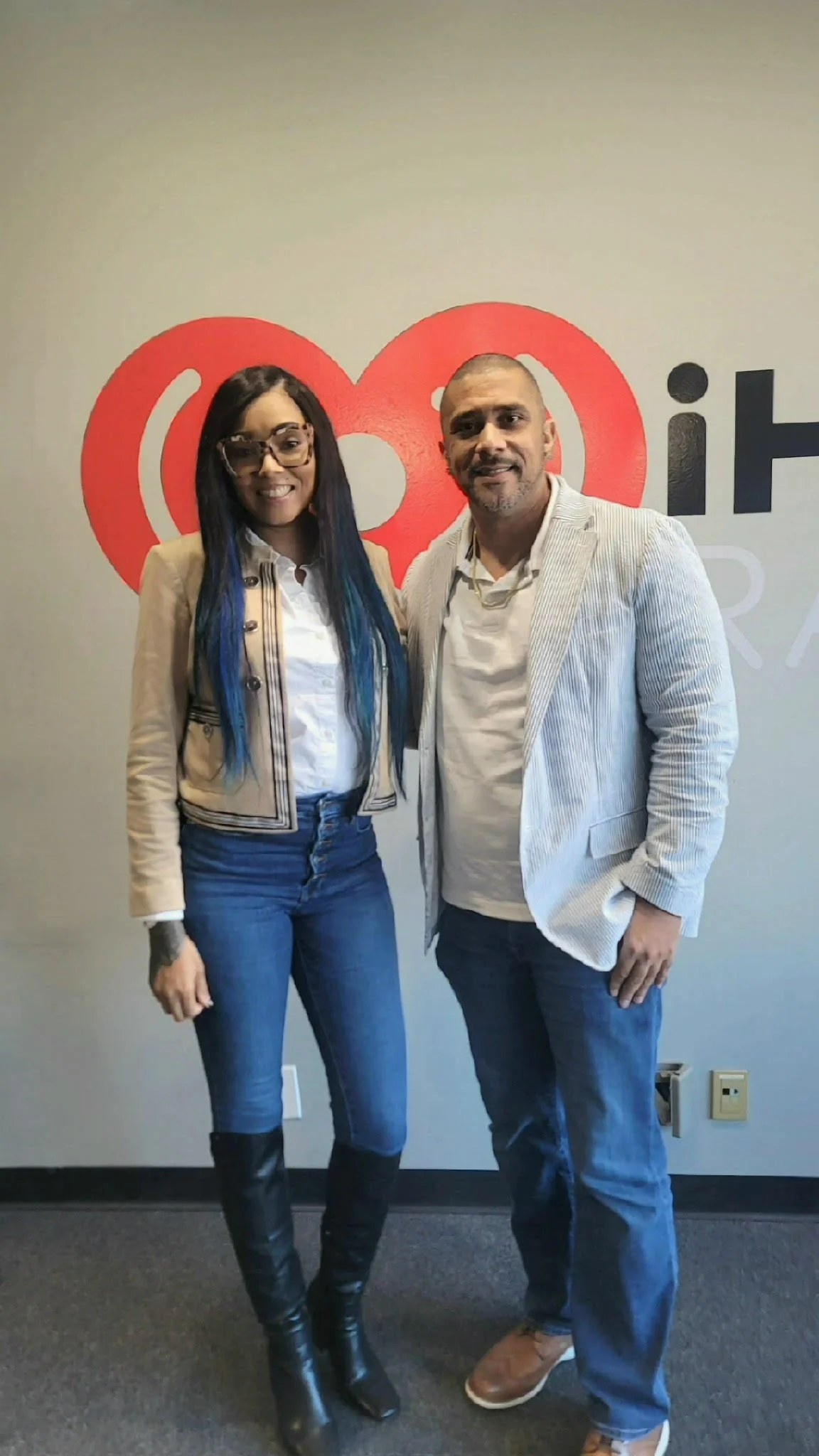 Two people standing together indoors in front of a wall with a large red and black logo. The woman on the left has long dark hair with blue highlights, glasses, and is wearing a beige jacket, white shirt, blue jeans, and black knee-high boots. The man on the right has short hair, a beard, and is wearing a light-colored blazer, white shirt, blue jeans, and brown shoes.