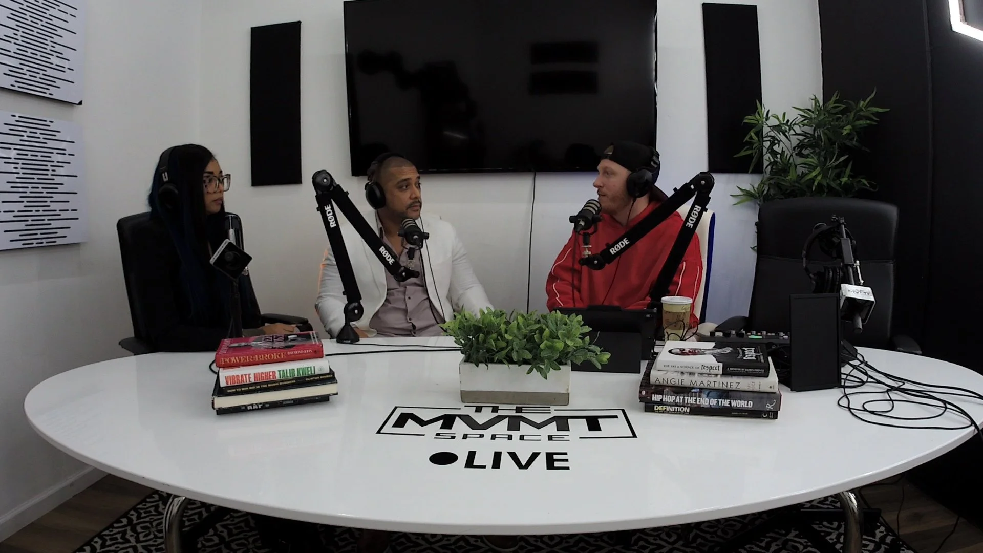 Three people sitting at a white table with microphones, books, and a plant, recording a podcast or radio show in a studio.