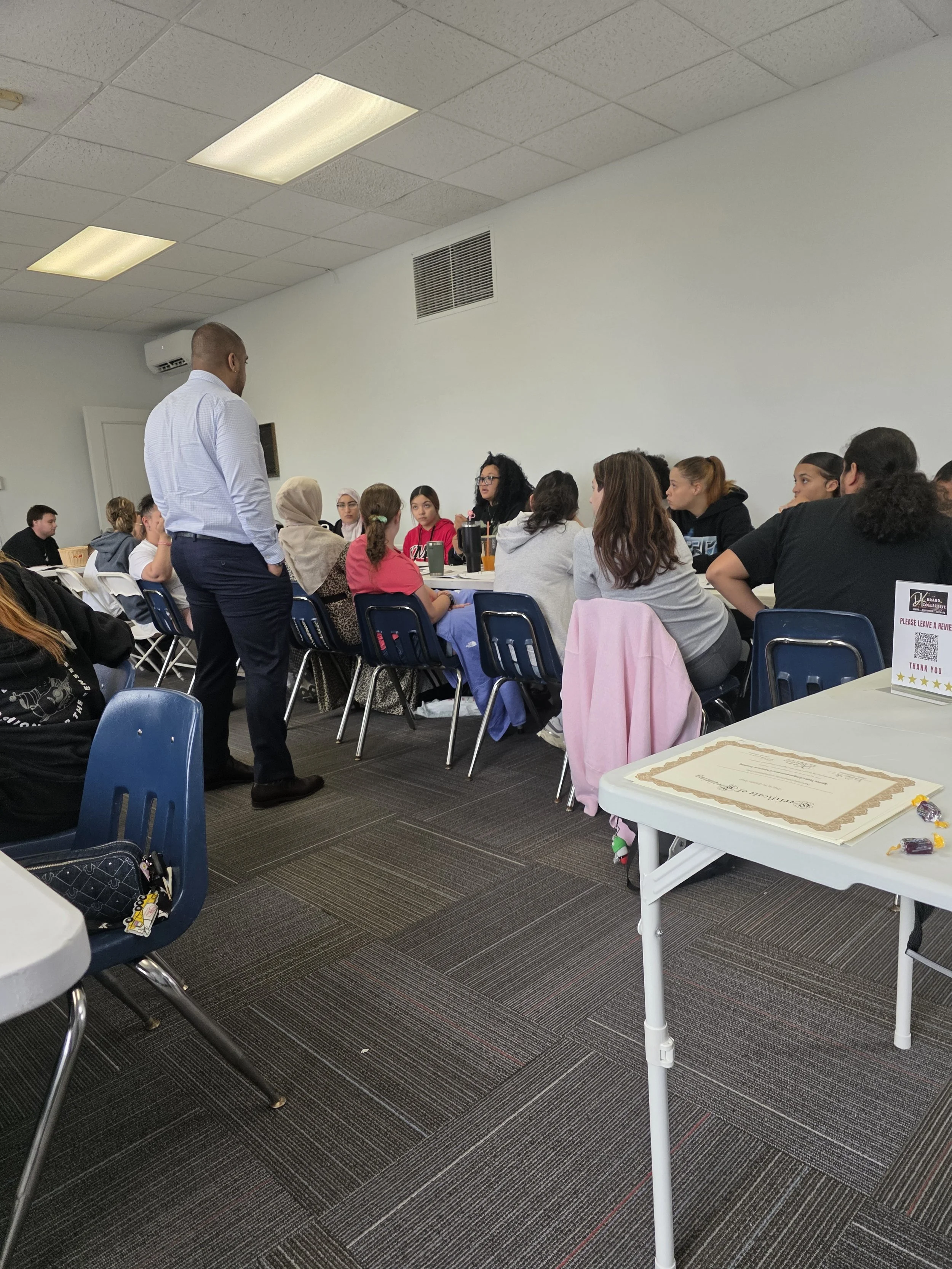 A man in a light blue shirt and dark pants stands at the front of a room with a group of women and girls seated at tables, listening to him speak. The room has white walls, a cubic ceiling, and a gray carpeted floor.