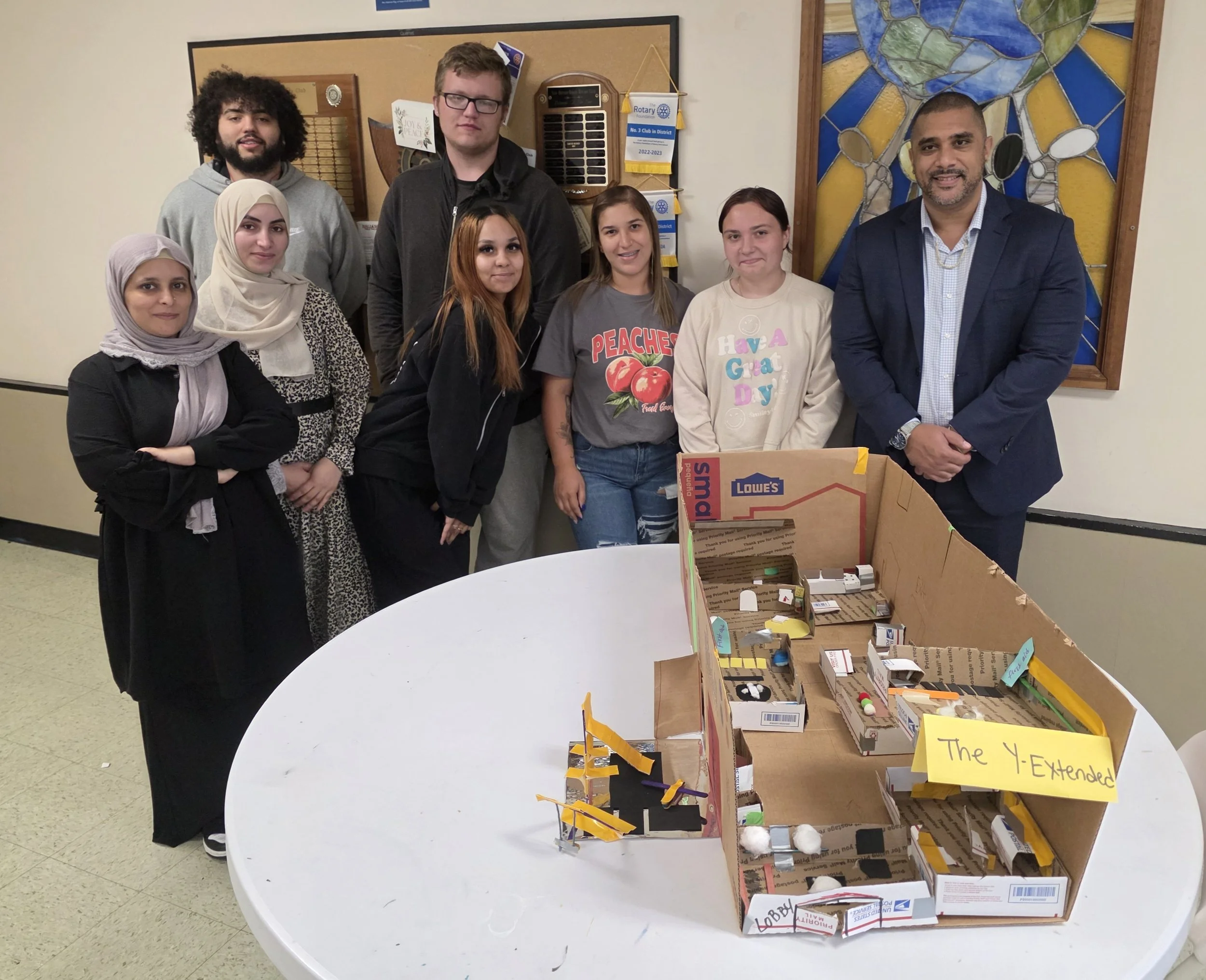 Group of nine people standing behind a table with a cardboard project. The group includes men and women, some wearing casual clothing, one woman in a hijab, and a man in a suit. The project displayed on the table has a sign that reads 'The Y-Extended' and appears to be a miniature model of a building or structure.