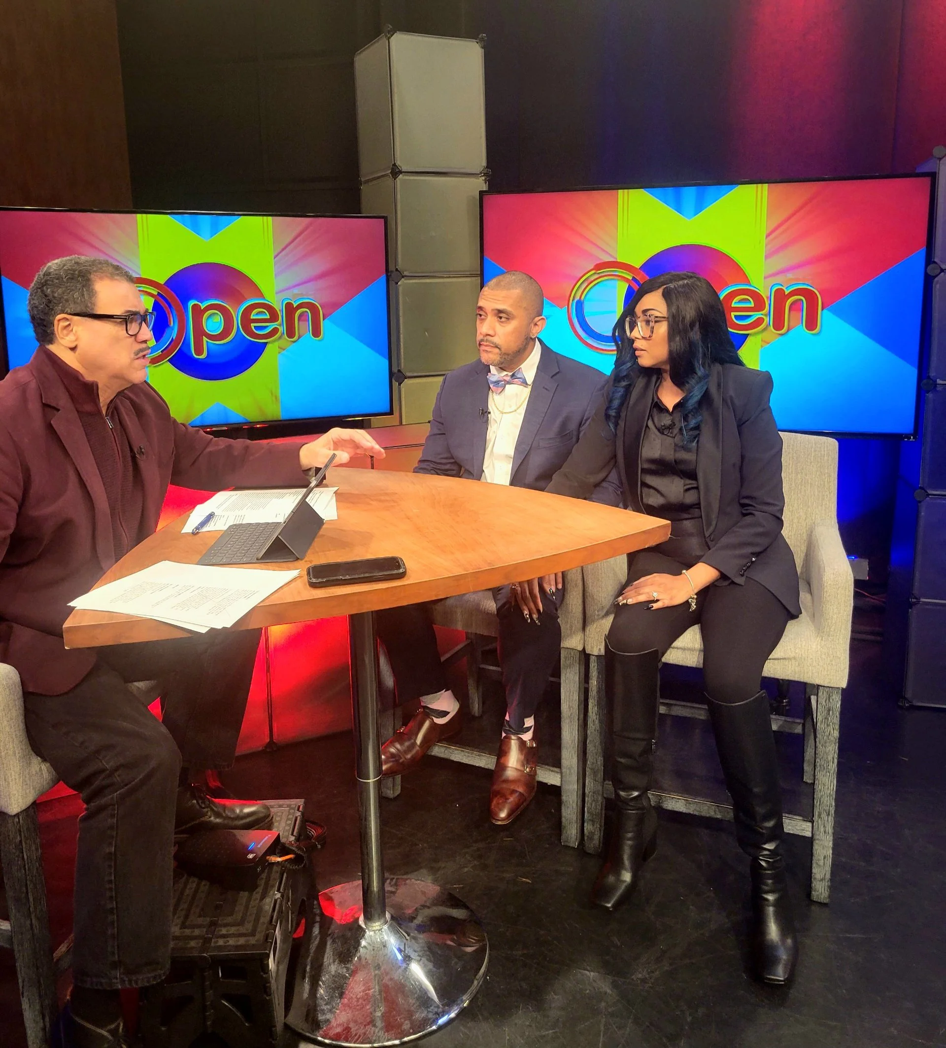 A television studio scene featuring three people engaged in discussion, with two TV screens displaying the word 'Open' in colorful graphics in the background.