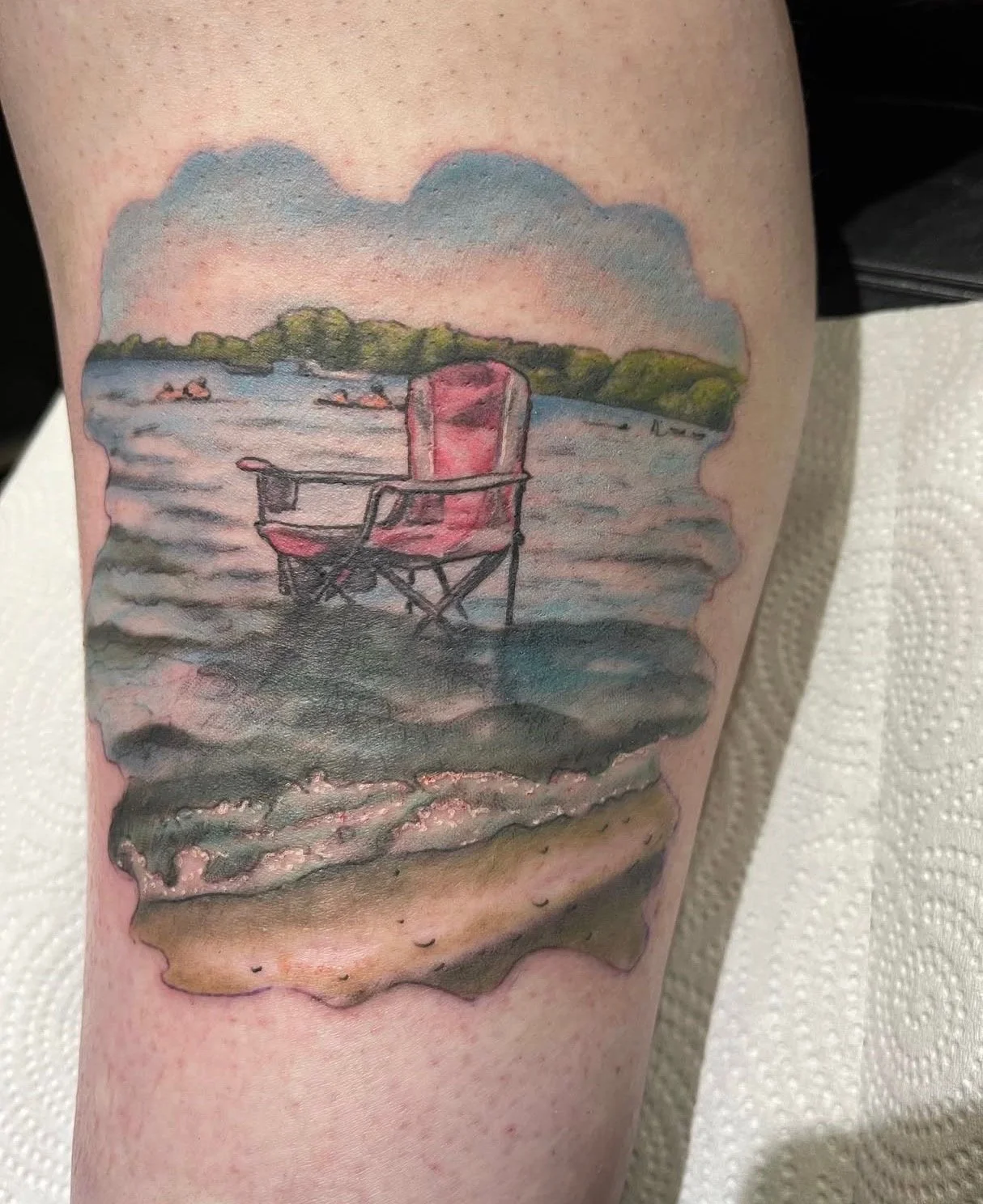 Sioux Falls South Dakota Tattoo Shop. Apex Ink. Tattoo of a beach scene with a pink camping chair in the water, with boats and trees in the background.