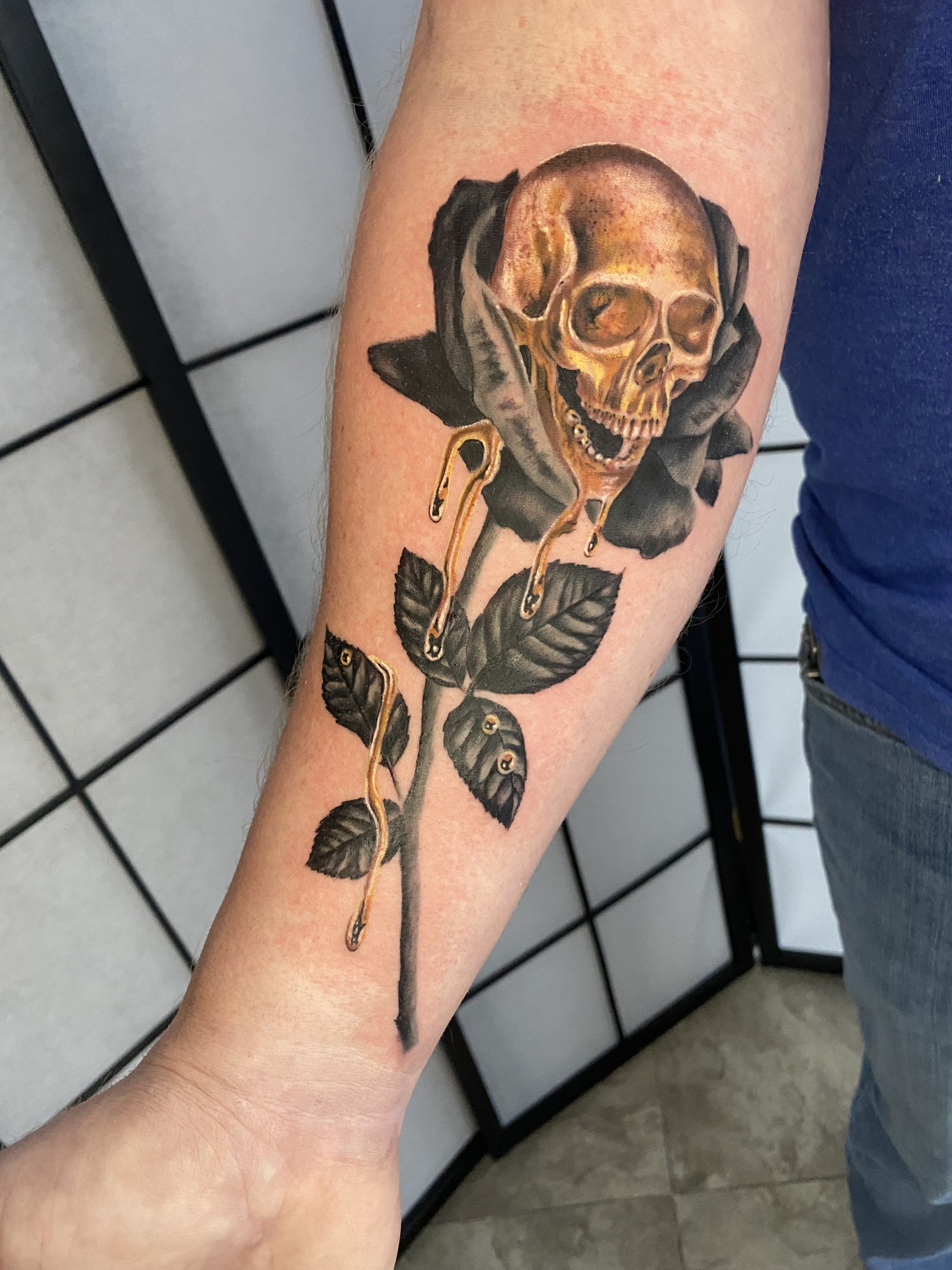 Tattoo of a human skull with gold accents, dripping like it's melting, attached to a black rose branch on the person's forearm.