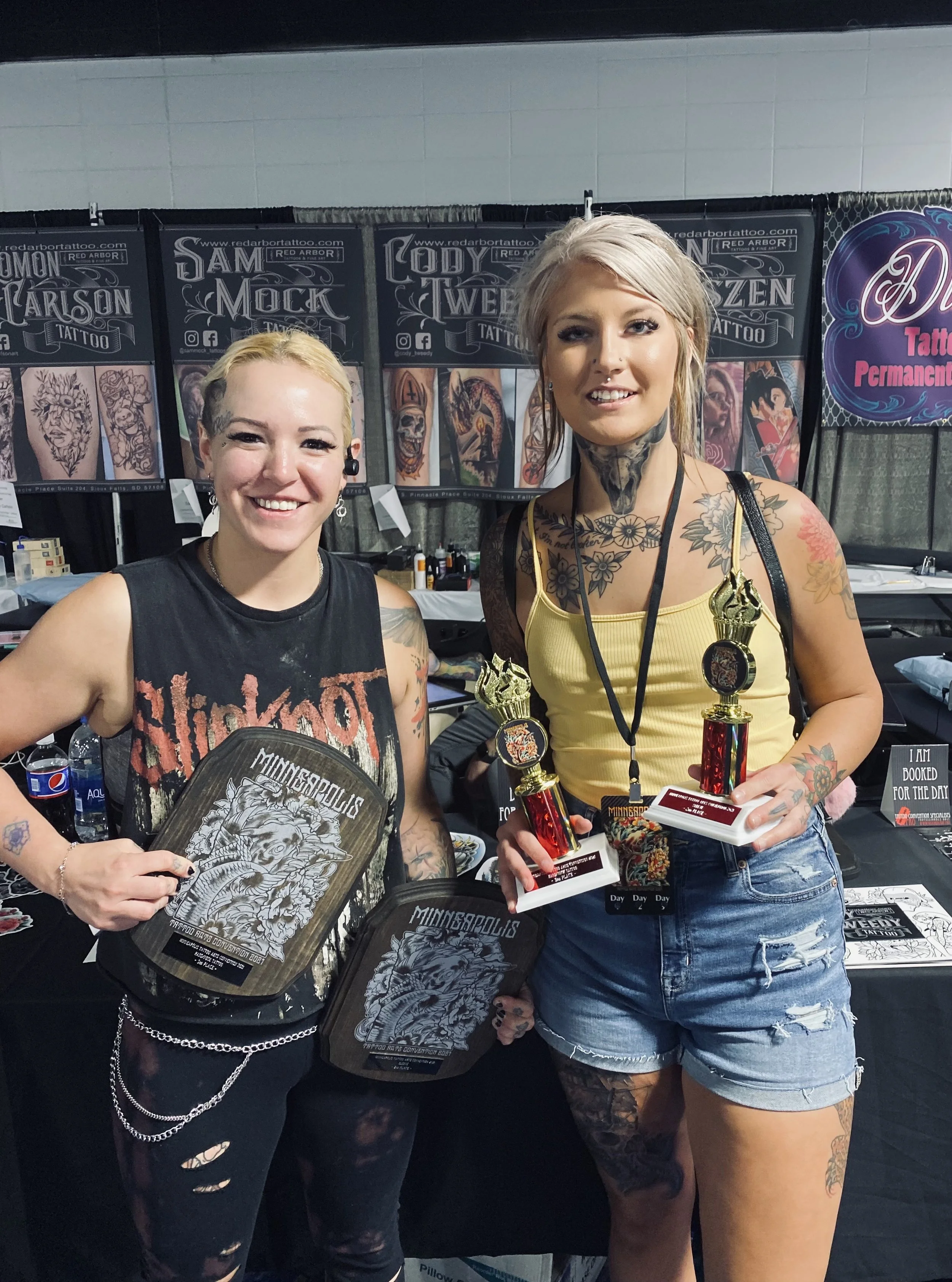 Two smiling women at a tattoo convention, holding trophies and merchandise, with tattoo booths in the background.
