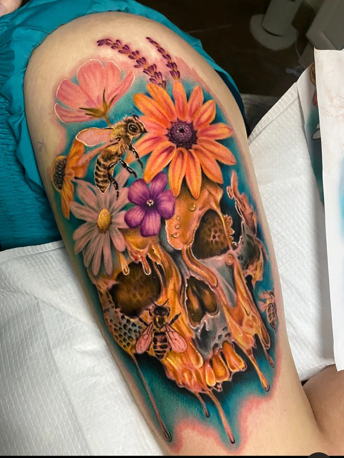 Colorful tattoo of a skull decorated with flowers and bees on a person's thigh.