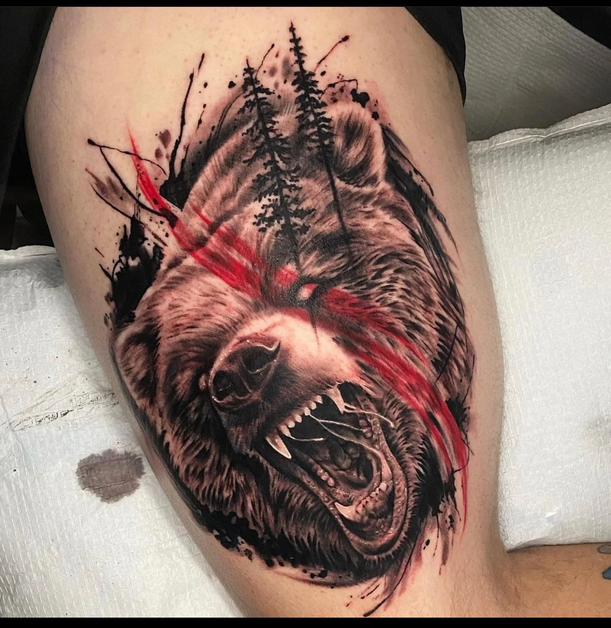 Tattoo of a fierce bear's head with an open mouth, showing teeth, surrounded by black ink splatters and two tall pine trees above, with red paint streaks across the face.