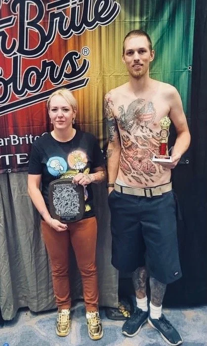 A man and woman stand side by side in front of a colorful banner at an event. The woman holds a plaque, and the man holds a trophy. The man is shirtless with numerous tattoos, including a large tattoo on his chest and legs showing an American flag and other designs.