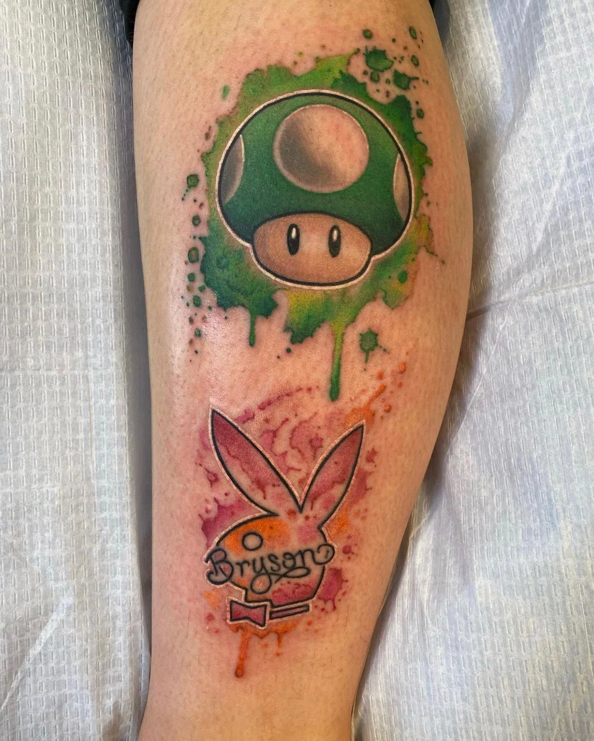 Tattoo of a Mario Bros. mushroom with green and dark shades, and the iconic Playboy bunny logo with the word 'Bruson' written inside it, featuring watercolor-style splashes of pink, orange, purple, and red.