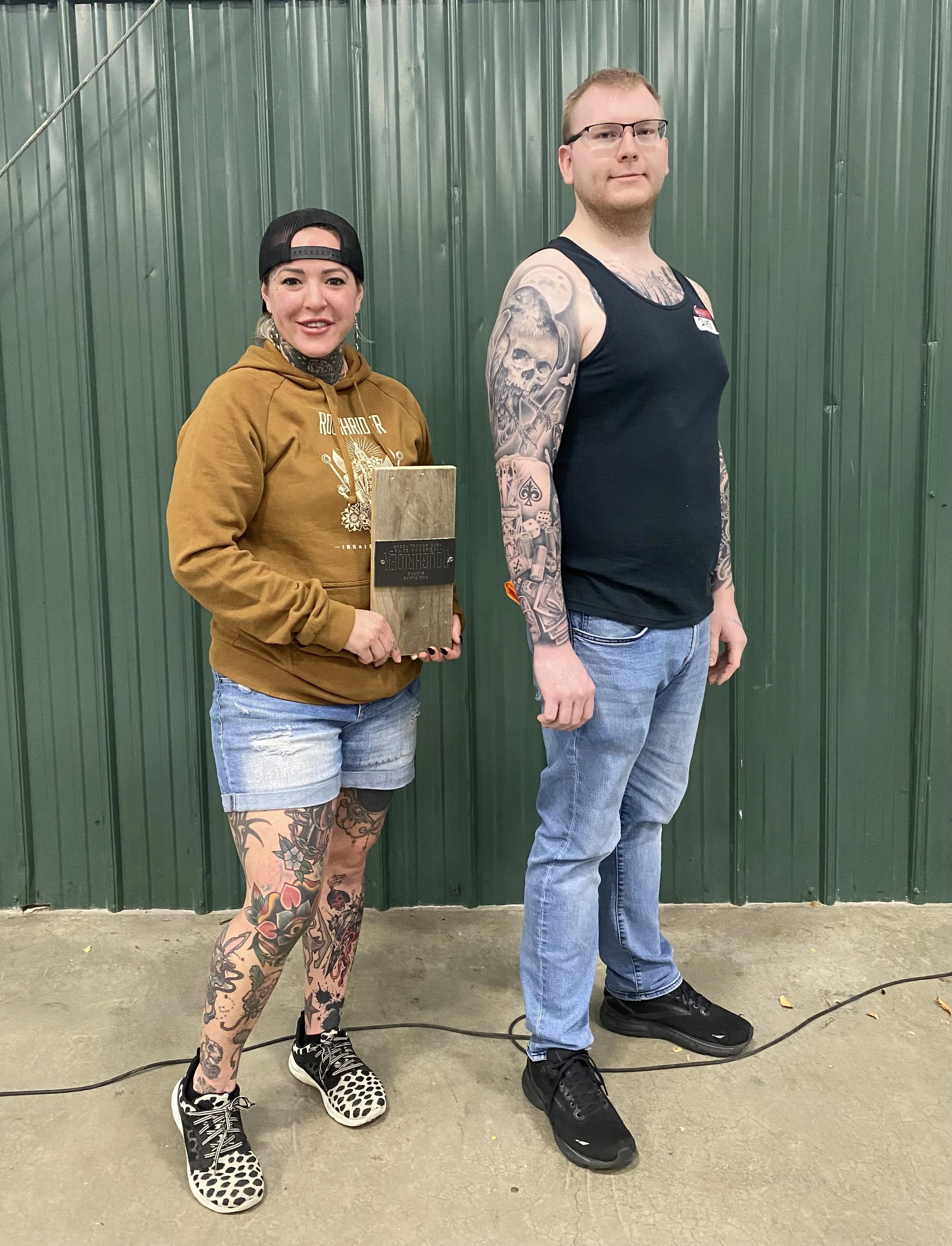A woman and a man standing indoors against a green metal wall. The woman is wearing a black cap, brown hoodie, denim shorts, and black and white patterned sneakers. She has tattoos on her legs and is holding a wooden plaque. The man is wearing glasses, a black tank top, blue jeans, and black shoes. He has tattoos on his arms.