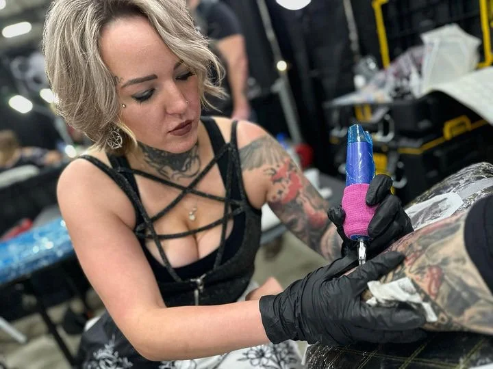 Female tattoo artist with blonde hair and tattoos on her arms, wearing black gloves, using a tattoo gun to ink a person on a table at a tattoo convention or studio.