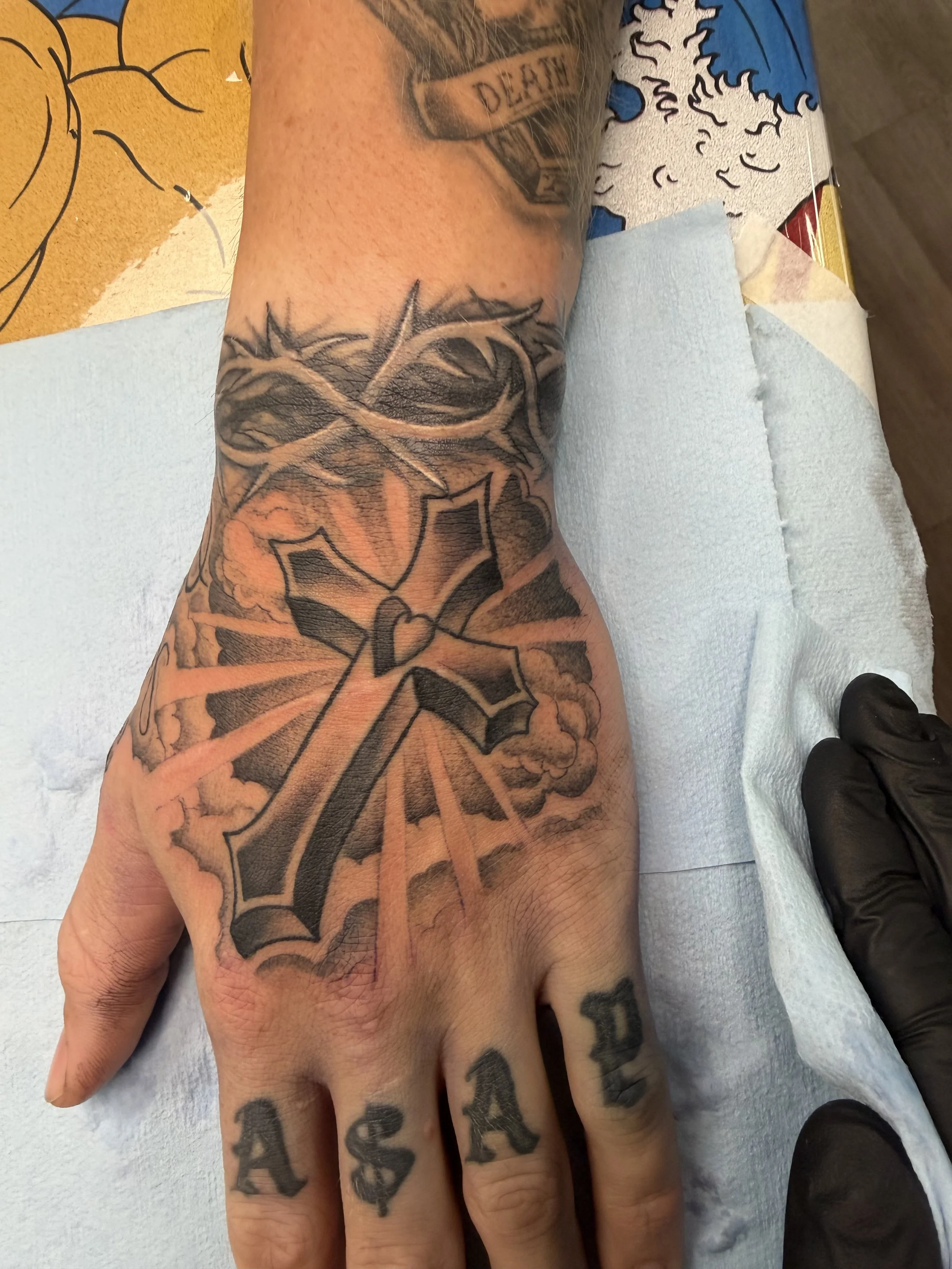 Sioux Falls South Dakota Tattoo Shop. Apex Ink. Tattoo of a cross surrounded by clouds on the hand, and a barbed wire design on the wrist. The person has tattooed letters 'A $ A E' on the fingers.