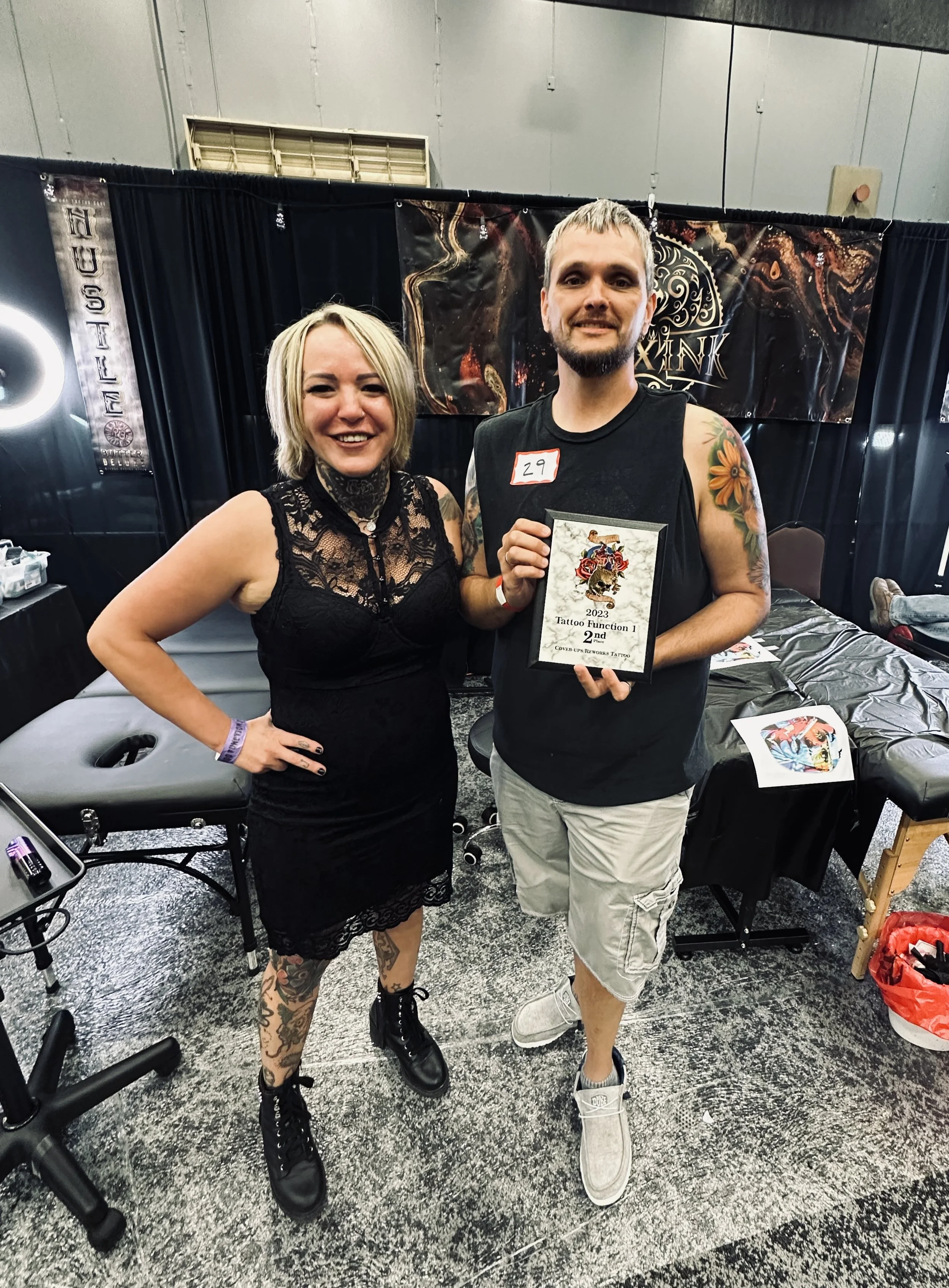 A woman in a black lace dress and tattoos is standing next to a man in casual clothing, both smiling and holding a framed award at a tattoo convention.