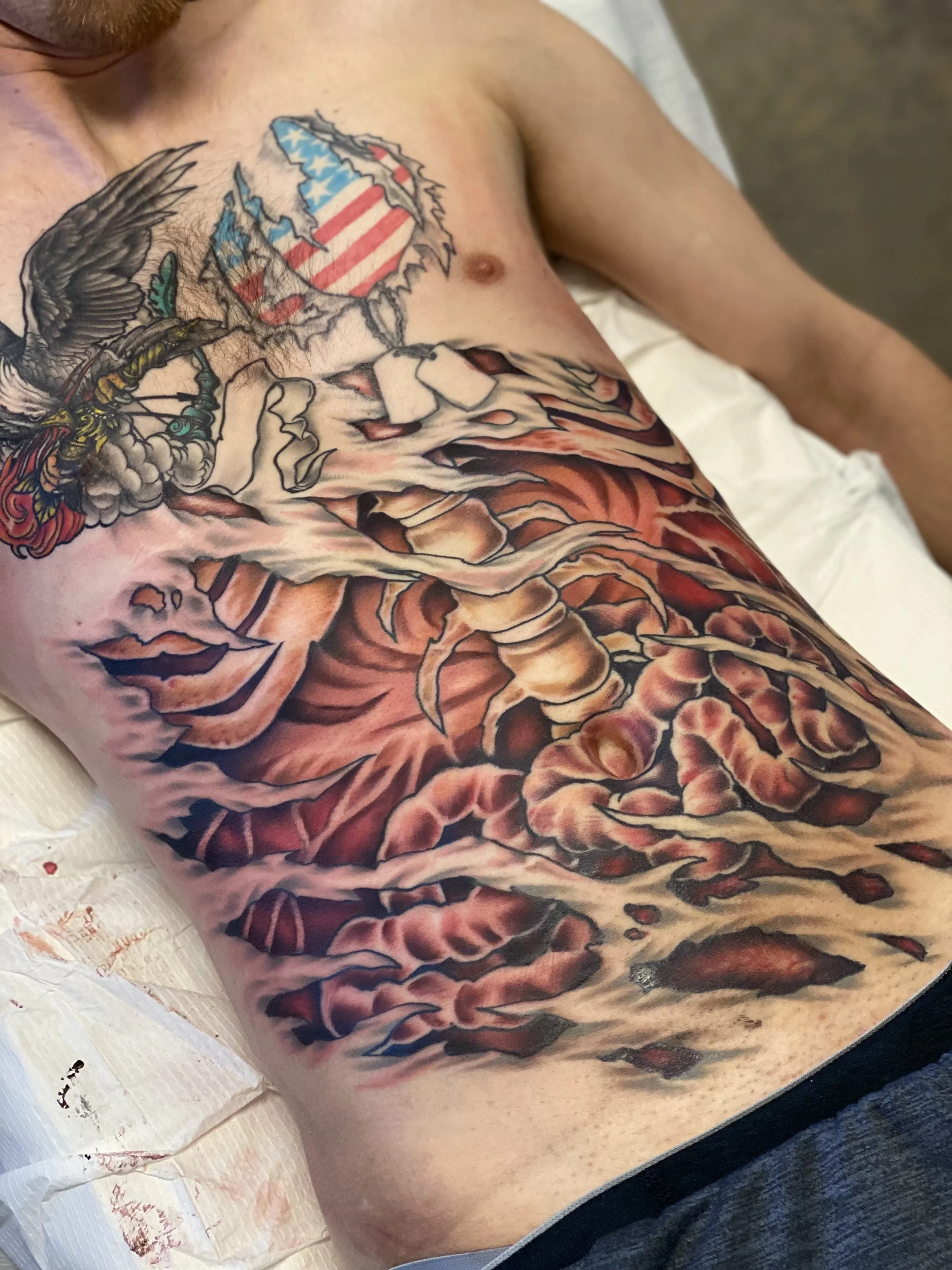 A man's torso with a large tattoo featuring an eagle, American flag, a woman's face, and abstract designs.